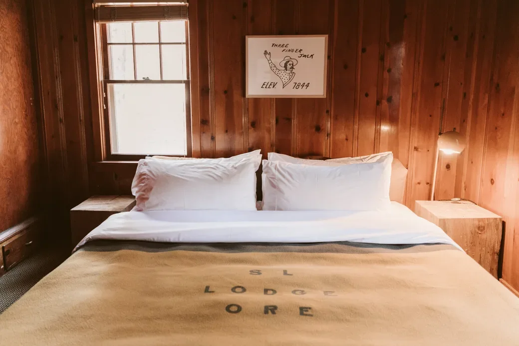 A cozy bedroom with wooden walls, a window, a bed with white pillows and sheets, and a beige blanket with 'S.L. Lodge ORE' printed on it. Above the bed hangs a framed drawing of a cartoon figure with the text 'Three Finger Jack,' 'Elev. 7844'.