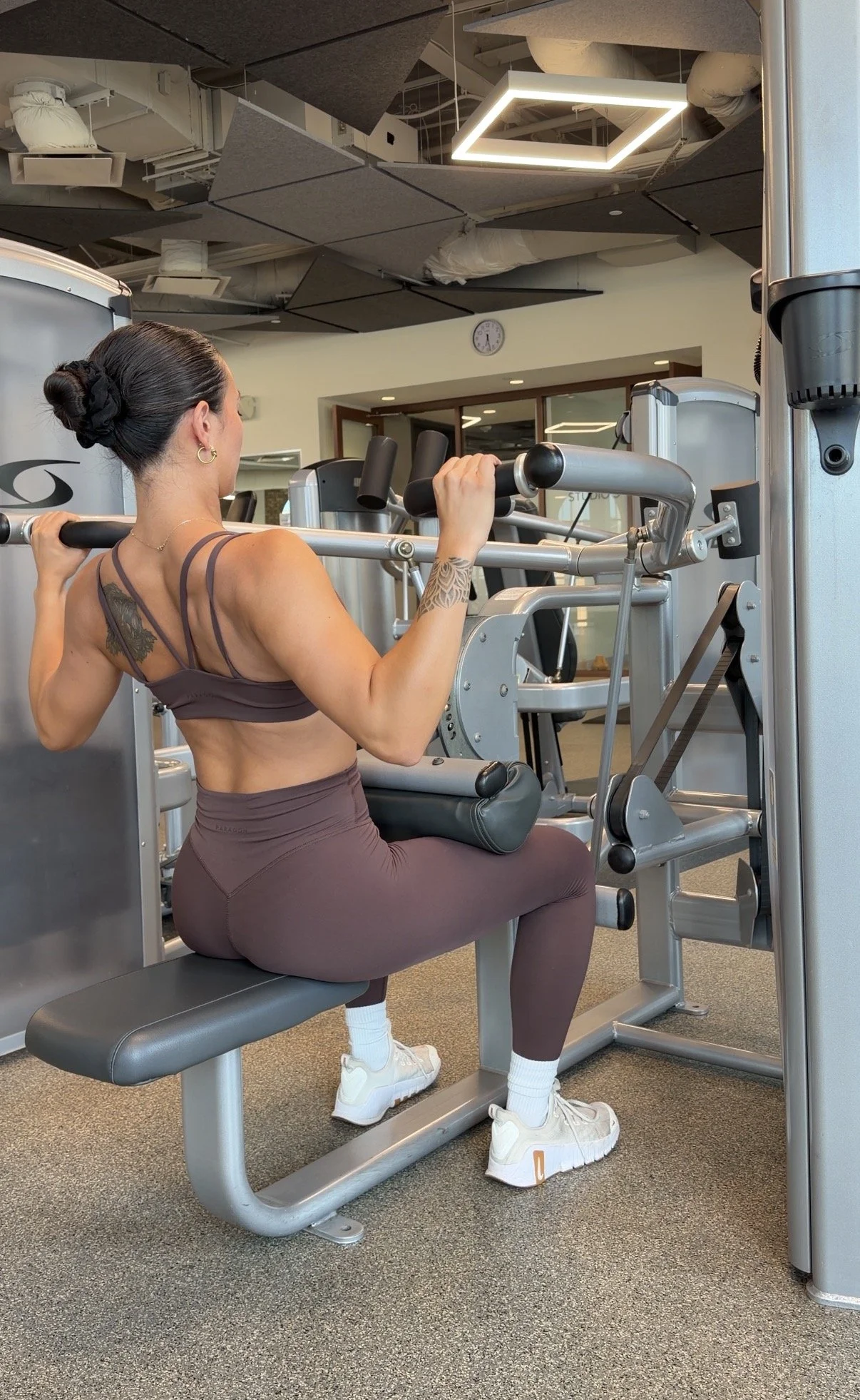 A woman with dark hair in a bun and tattoos on her shoulder and arm is seated on a gym exercise machine, lifting a barbell across her shoulders. She wears a brown sports bra and matching leggings, white sneakers, and white socks. The gym has a gray carpeted floor, adjustable lighting, and a ceiling with exposed ductwork.