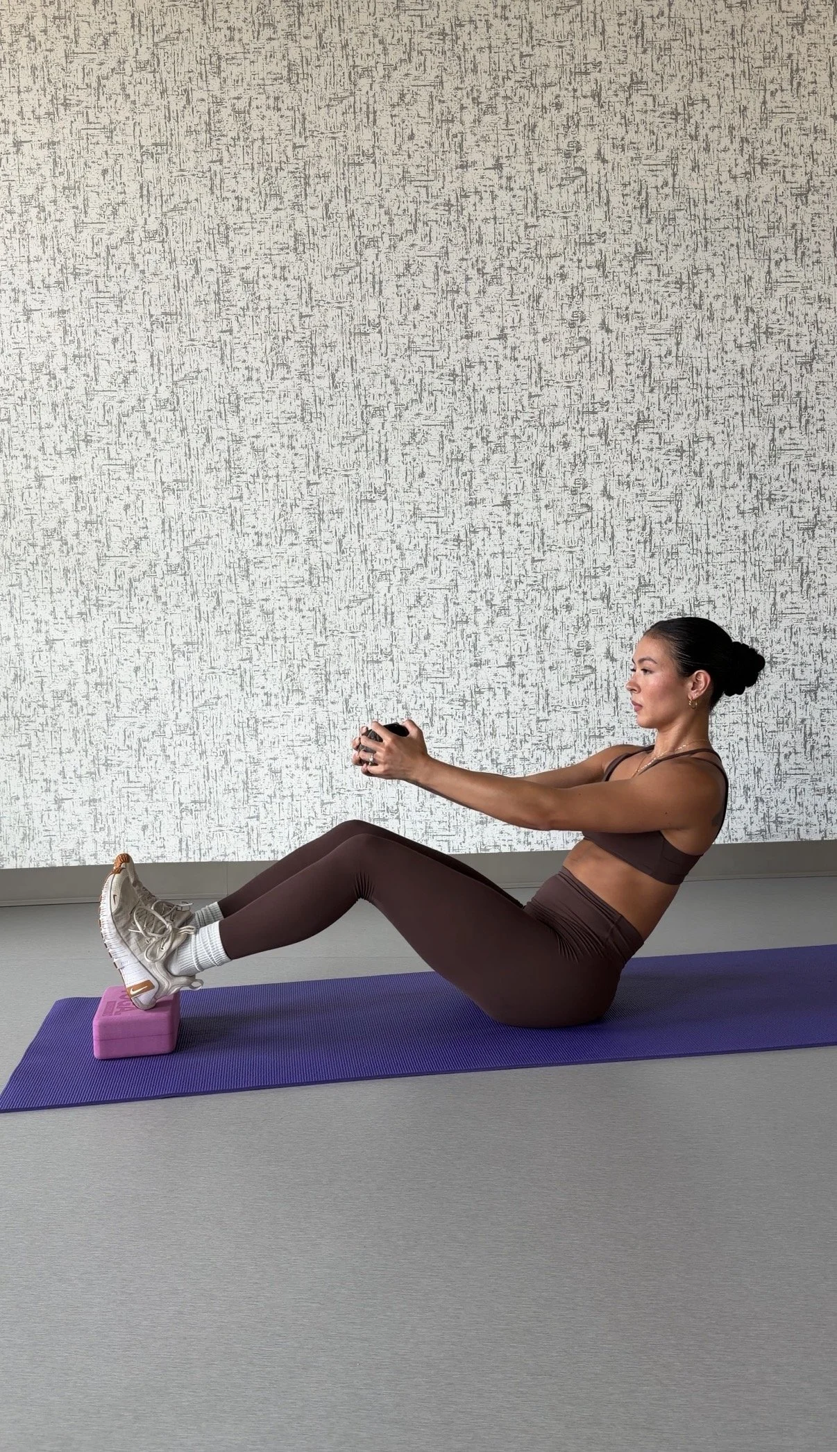 Woman exercising indoors, sitting on a purple yoga mat with her legs extended and feet resting on a pink block, holding a small weight with both hands. She is wearing a brown sports bra, matching leggings, and white sneakers with tan accents. The background features a textured gray wall.