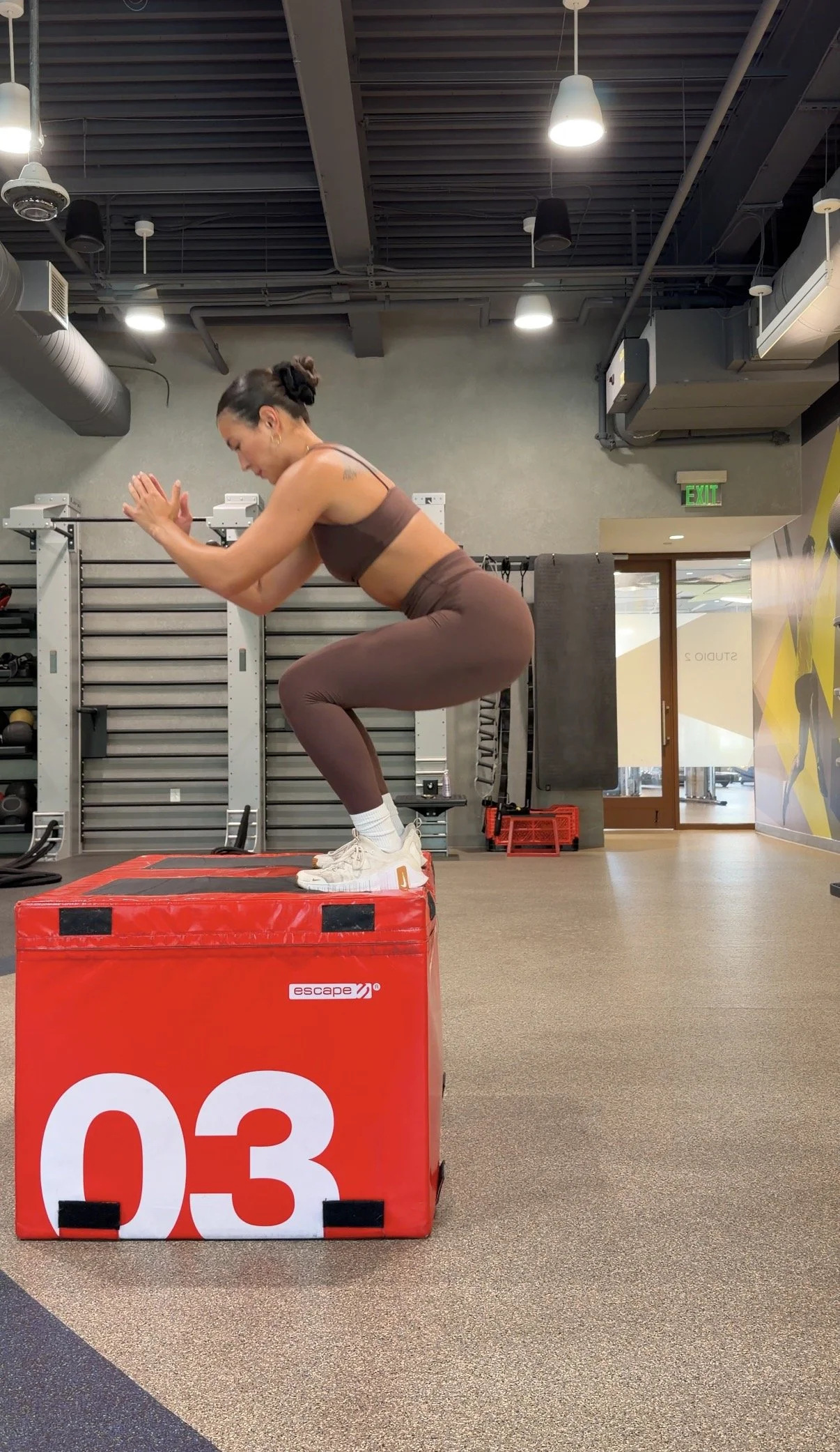 A woman wearing workout clothes is at a gym, standing on a red padded box, with her hands pressed together and her knees slightly bent, in a squat exercise. The gym has a modern industrial look with exposed ceiling pipes and hanging lights.