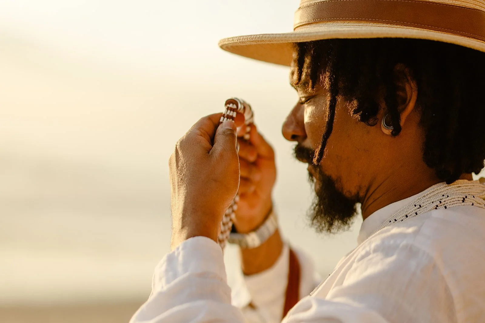 A man with a hat and earrings holds prayer beads close to his face, eyes closed, during a serene moment outdoors with a blurred person in the background.