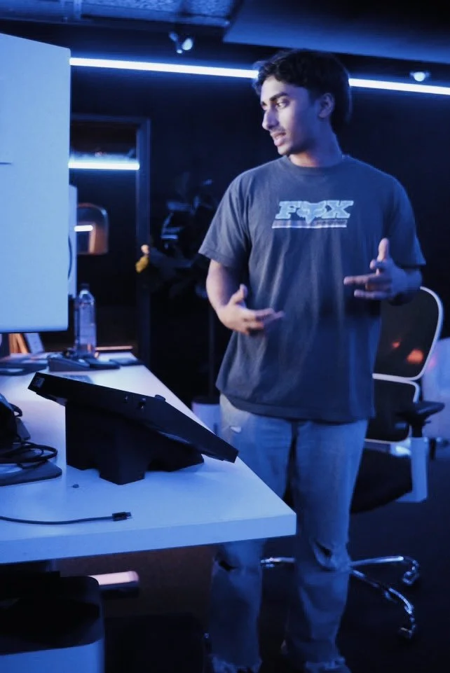 A young man with dark hair wearing a gray Fox Racing t-shirt in a dark room with blue lighting, standing at a desk with a computer monitor.