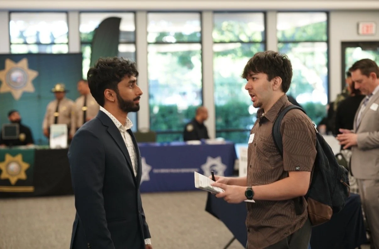 Two men in conversation at a police event or conference, with police officers and booths in the background.