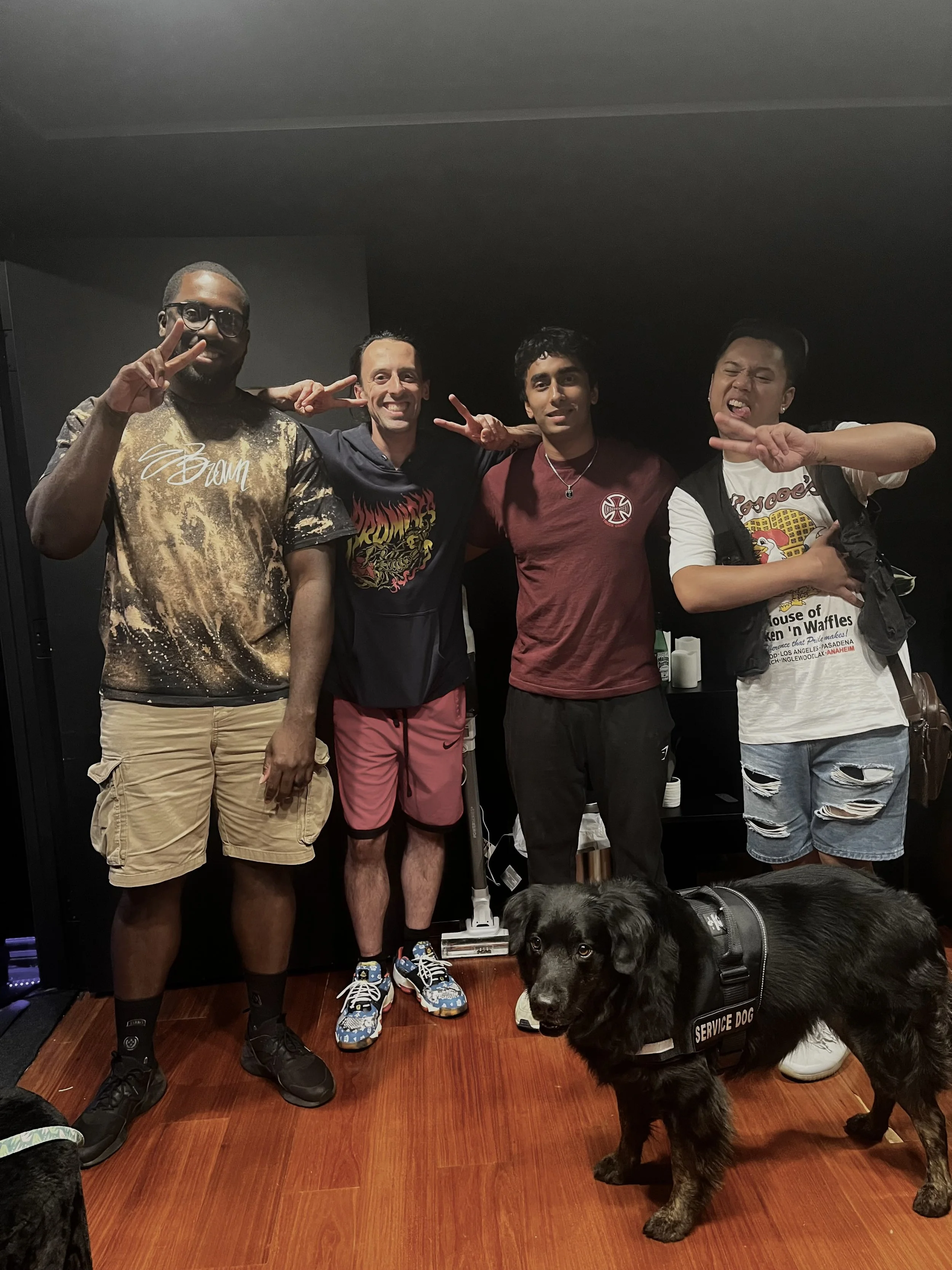 Four men and a service dog in a room with dark walls and wooden floor. The men are posing for the photo, with some making peace signs. The service dog, a black Labrador, is in front, wearing a vest labeled 'SERVICE DOG' and looking at the camera.
