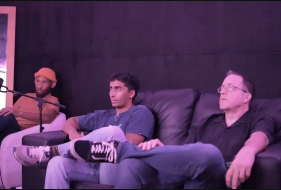 Three men sitting on a black leather couch in a dark room, appearing to listen attentively, with one man wearing a green shirt and sneakers, another in a black shirt, and the third in an orange hat and shirt.