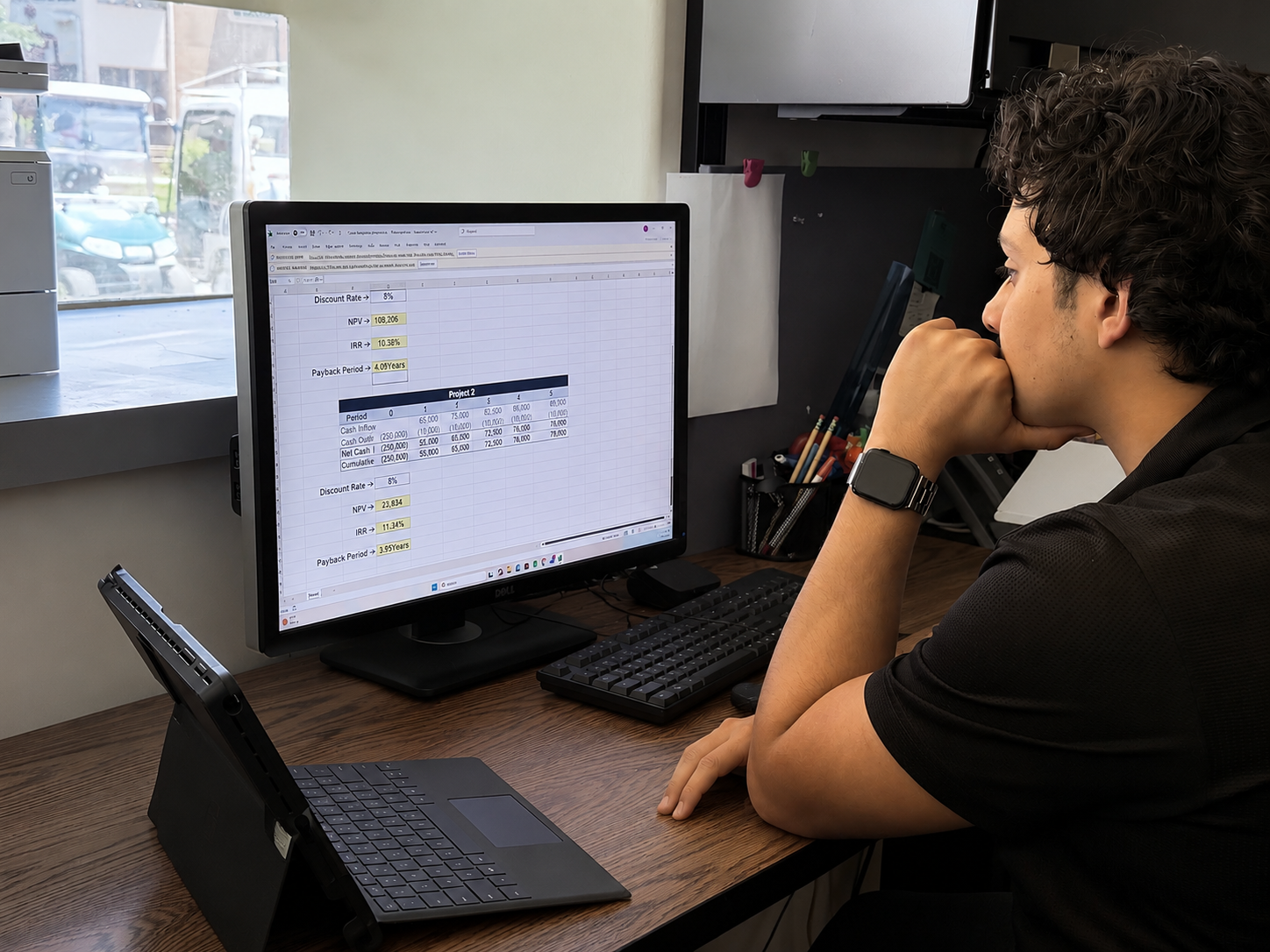 Young man working on financial spreadsheets on a desktop computer at a wooden desk, with a tablet on a stand, in an office with a window showing parked cars outside.