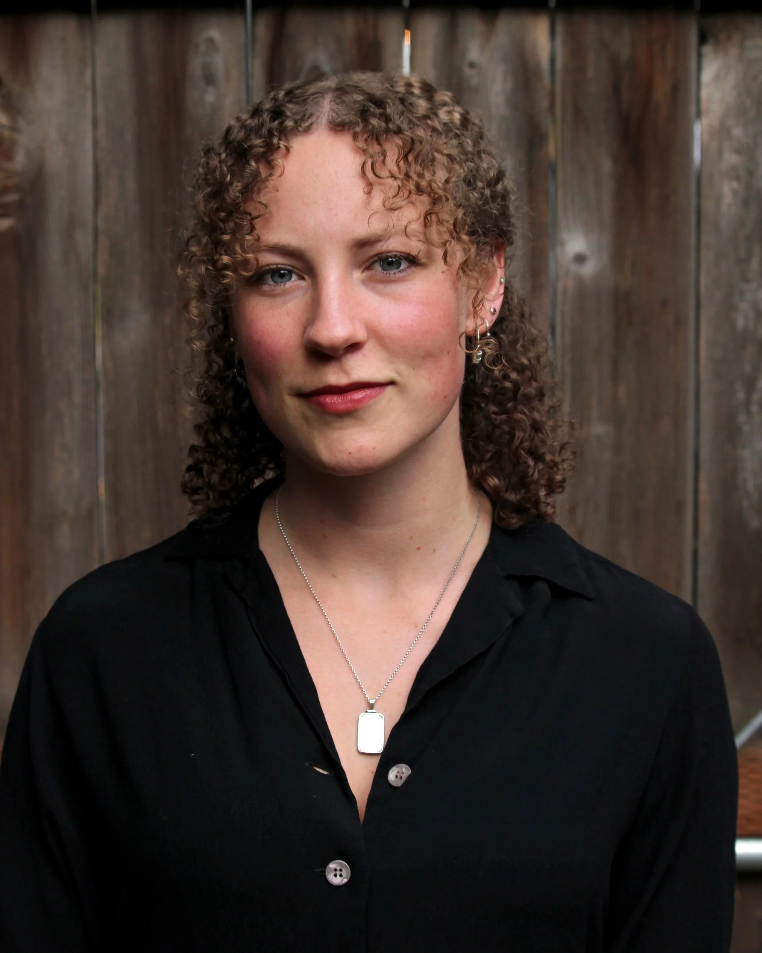 A woman with curly brown hair wearing a black top and silver necklace, standing in front of a wooden wall.