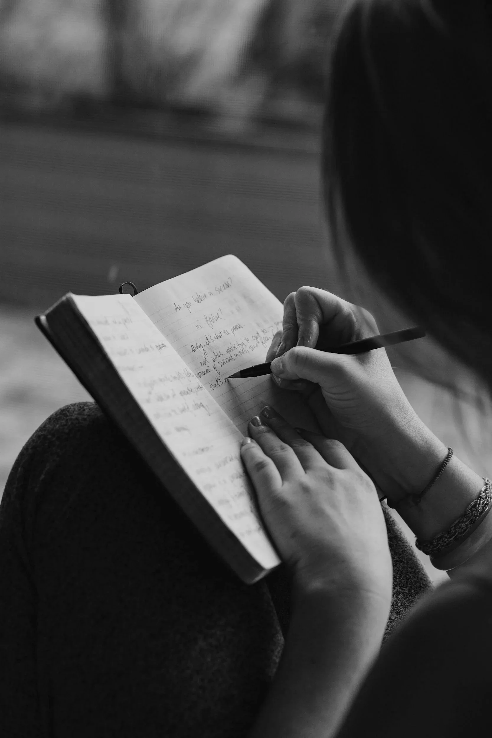 A person holding a notebook and writing with a pen, sitting with legs crossed on a textured surface, in black and white.