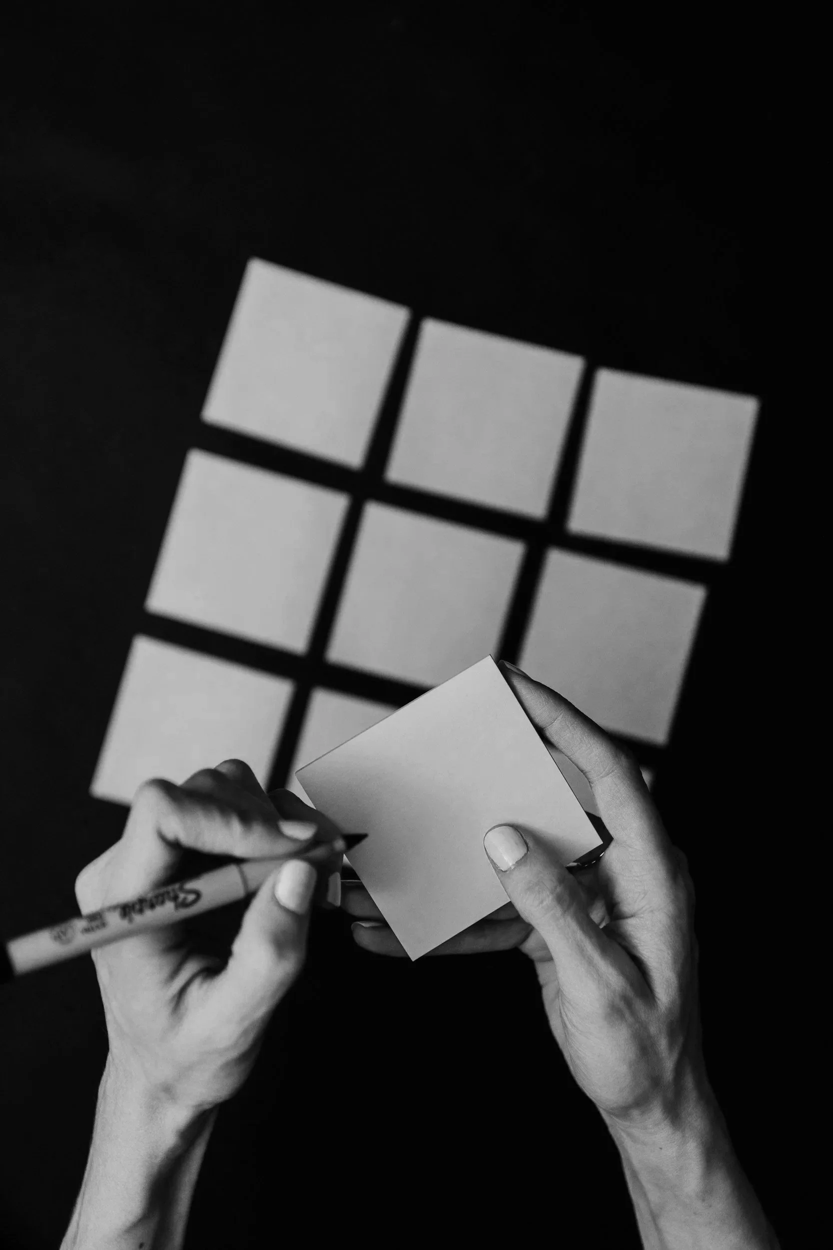 Hands writing on a small notebook with a black marker, with a grid of light-colored squares casting shadows on a dark background.