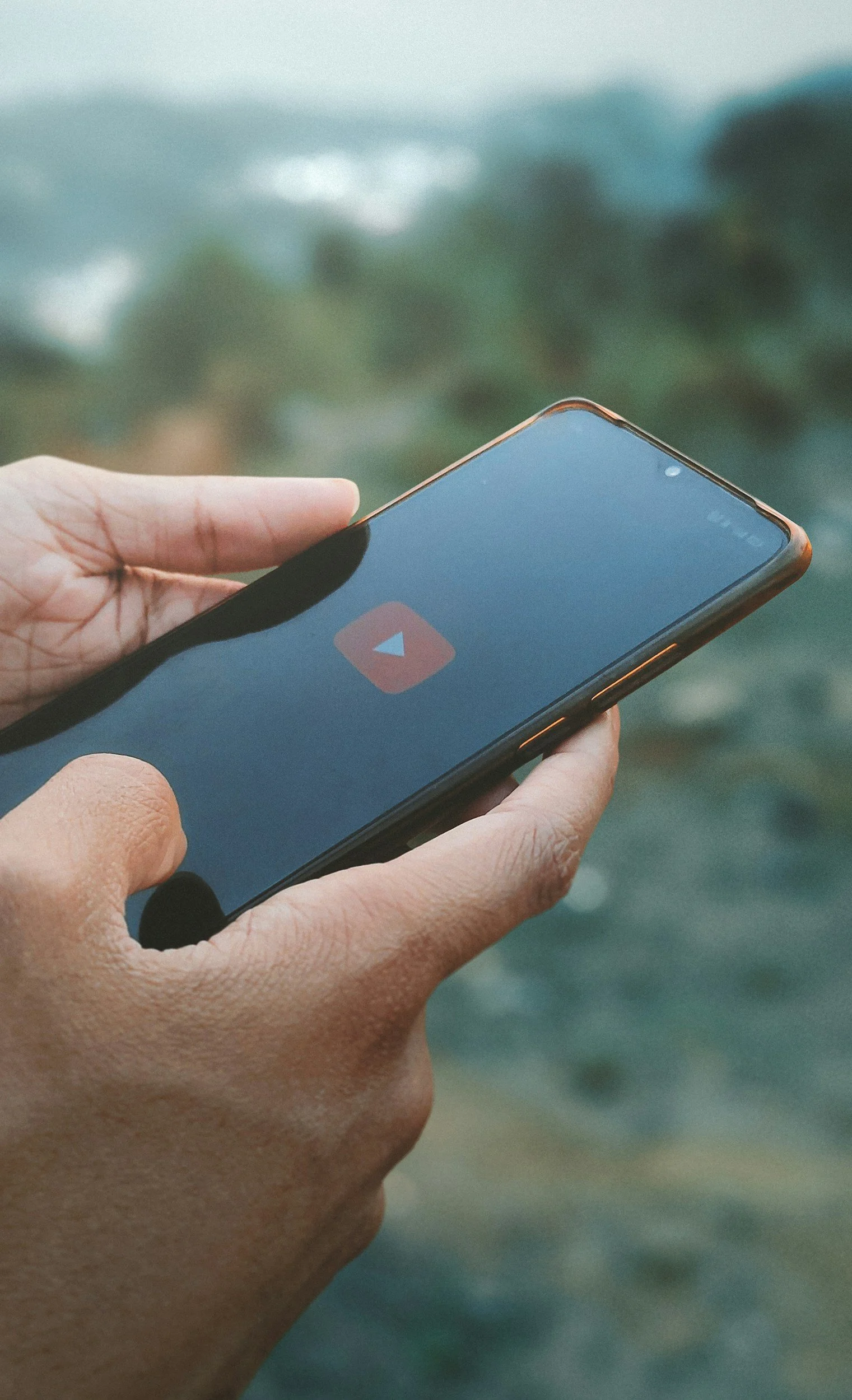Close-up of a person's hand holding a smartphone with the YouTube app open on the screen, against a blurred green and blue outdoor background.