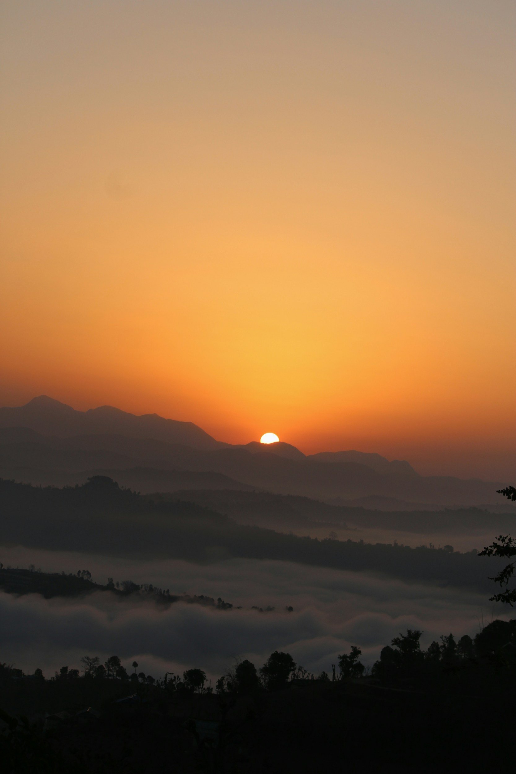 Sunset over mountains with layered hills, some trees, and fog in the valleys, creating a tranquil landscape.