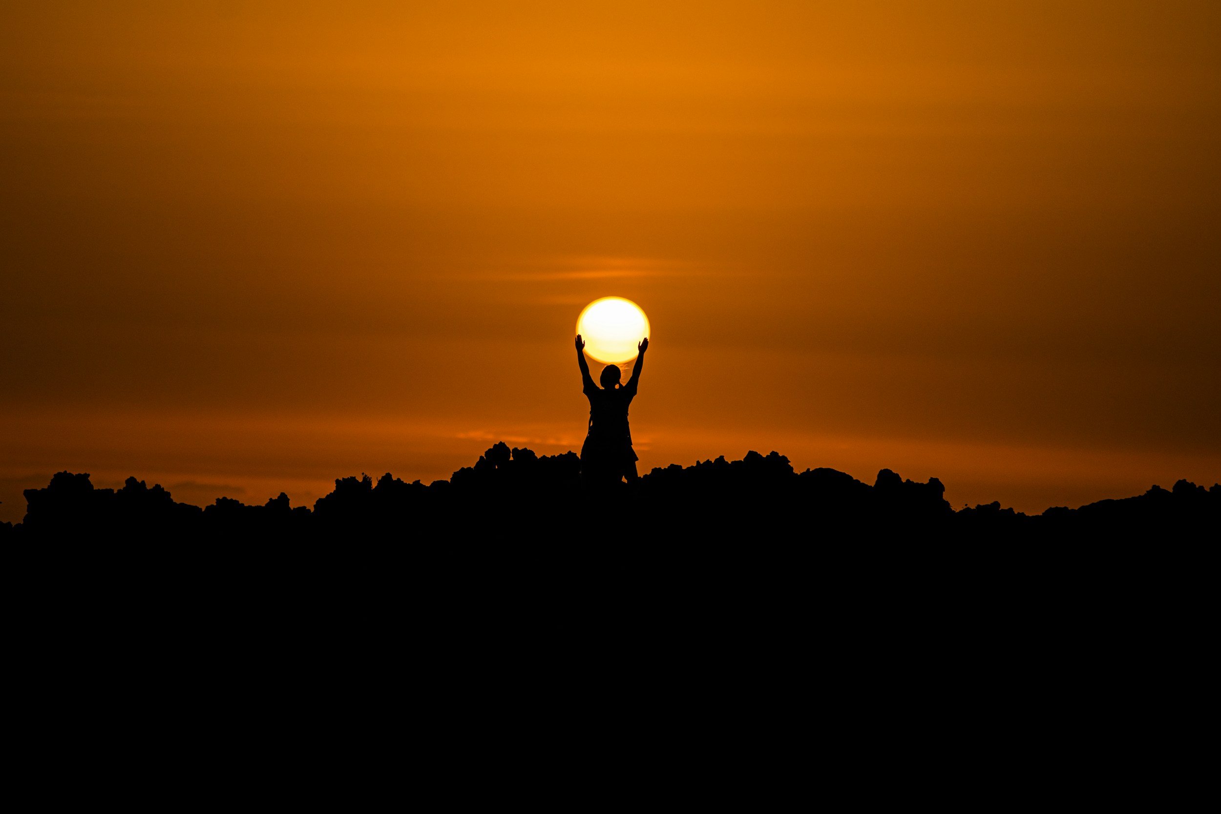 A person standing outdoors at sunset with their arms raised, holding the sun as if they are lifting it. The sky is orange with some clouds, and there are dark silhouettes of rocks or trees in the foreground.