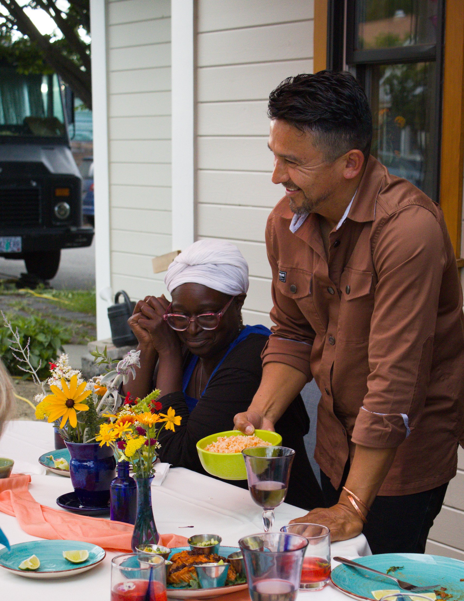 A man with dark hair and a beard, dressed in a brown shirt, laughs as he stands next to a woman with dark skin and a white head covering, sitting at a table with food and flowers, outdoors near a house with beige siding.