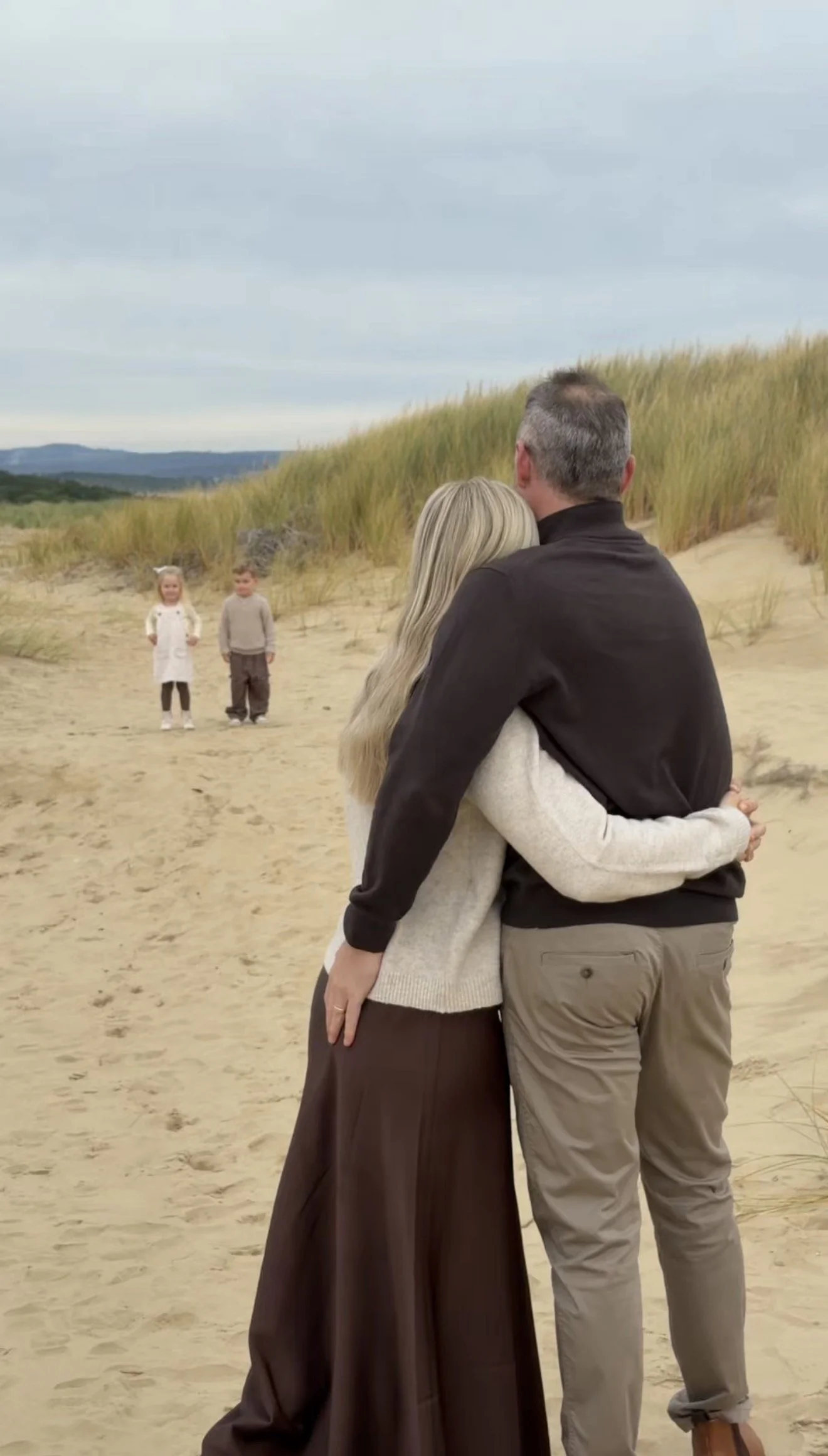 A couple hugging on the beach with two children walking in the background.