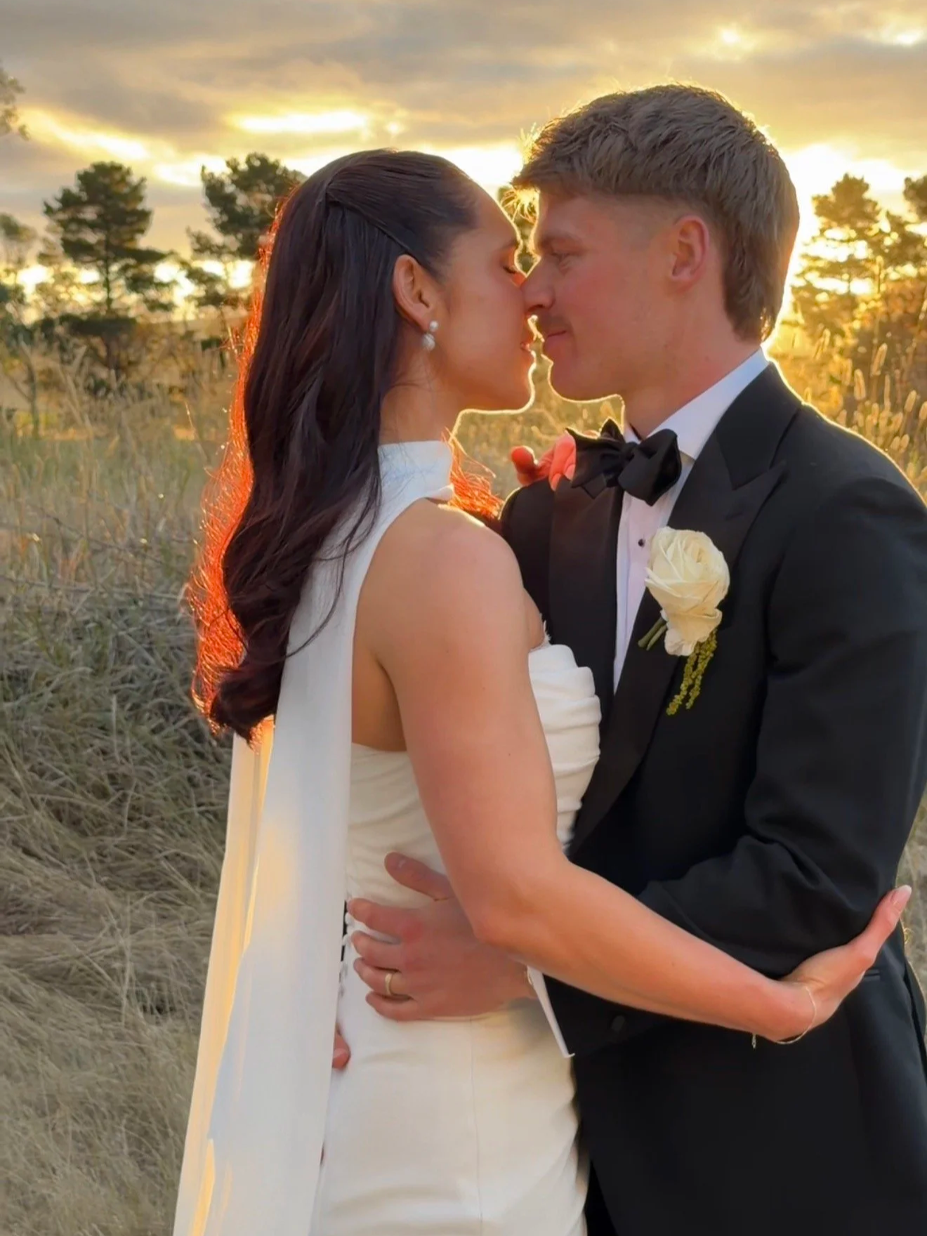 A newlywed couple, a woman in a white wedding dress and a man in a tuxedo, embracing and about to kiss in an outdoor setting at sunset, with trees and a partly cloudy sky in the background.