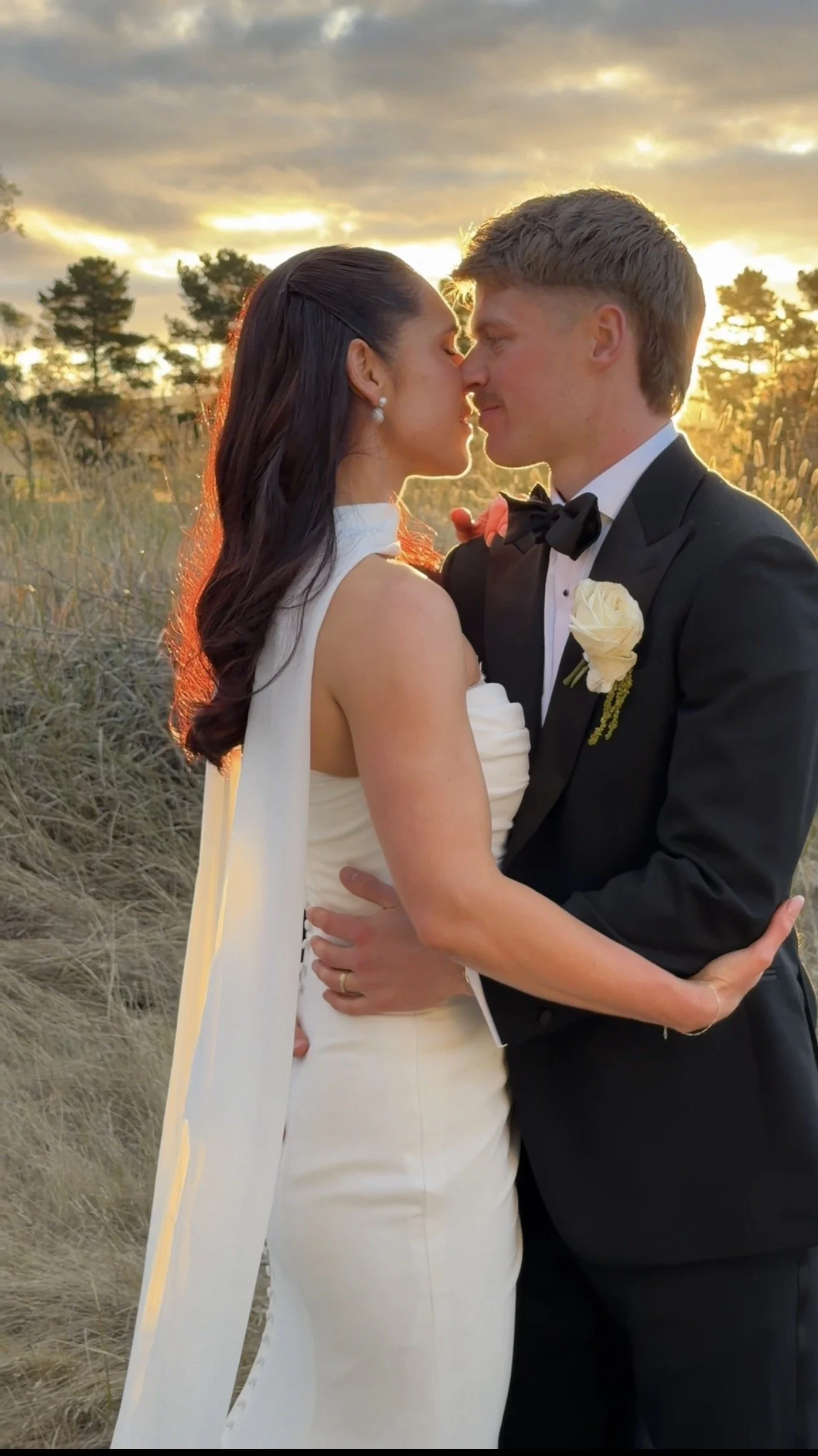 A bride and groom sharing a kiss outdoors during sunset, with trees and sky in the background, the bride in a white wedding dress and the groom in a tuxedo with a boutonnière.