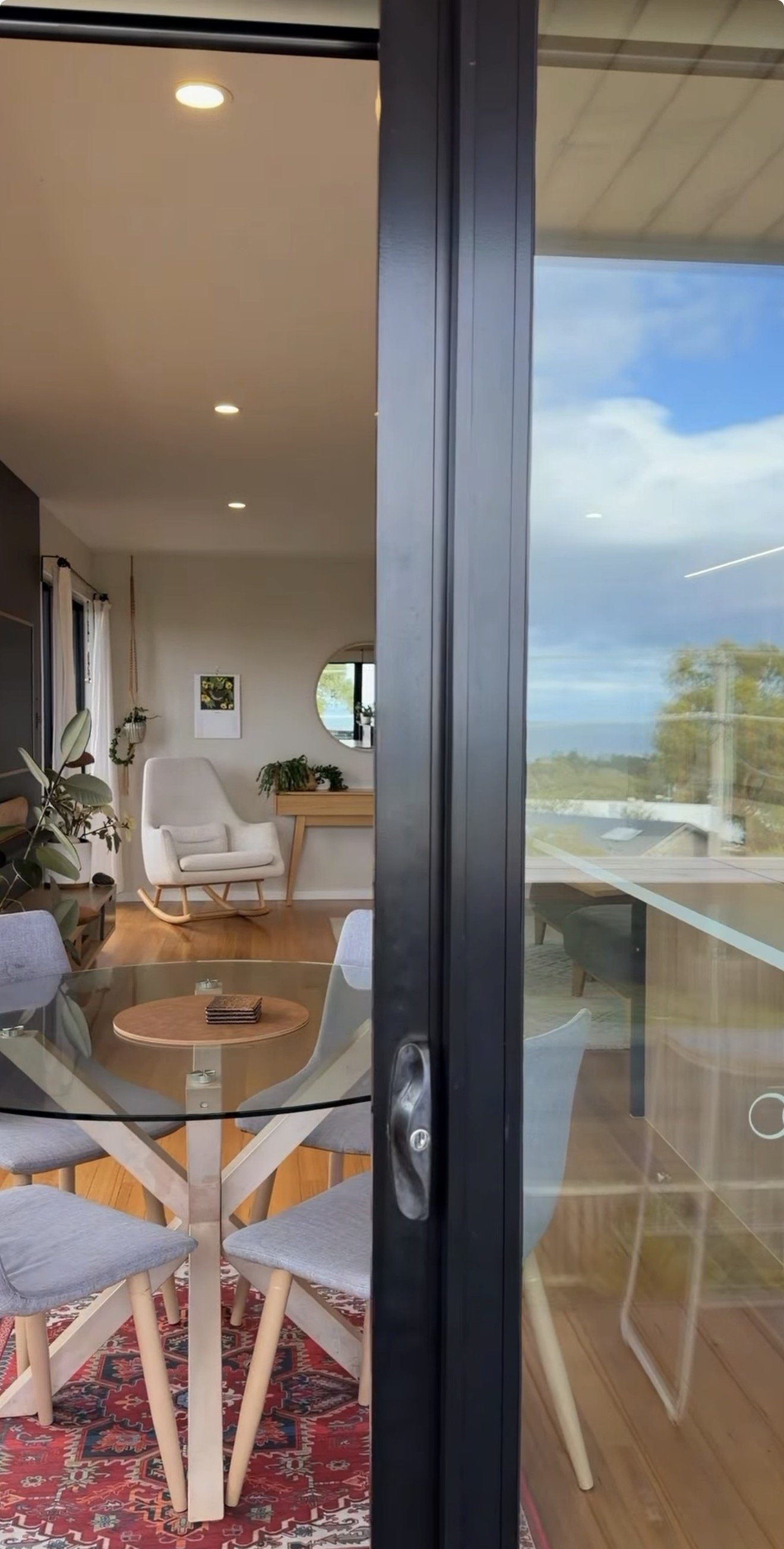 Living room and dining area seen through a glass door; interior includes a round glass table with four gray upholstered chairs, a white rocking chair, a wooden side table, potted plants, and artwork on the wall, with a view of blue sky and trees outside.