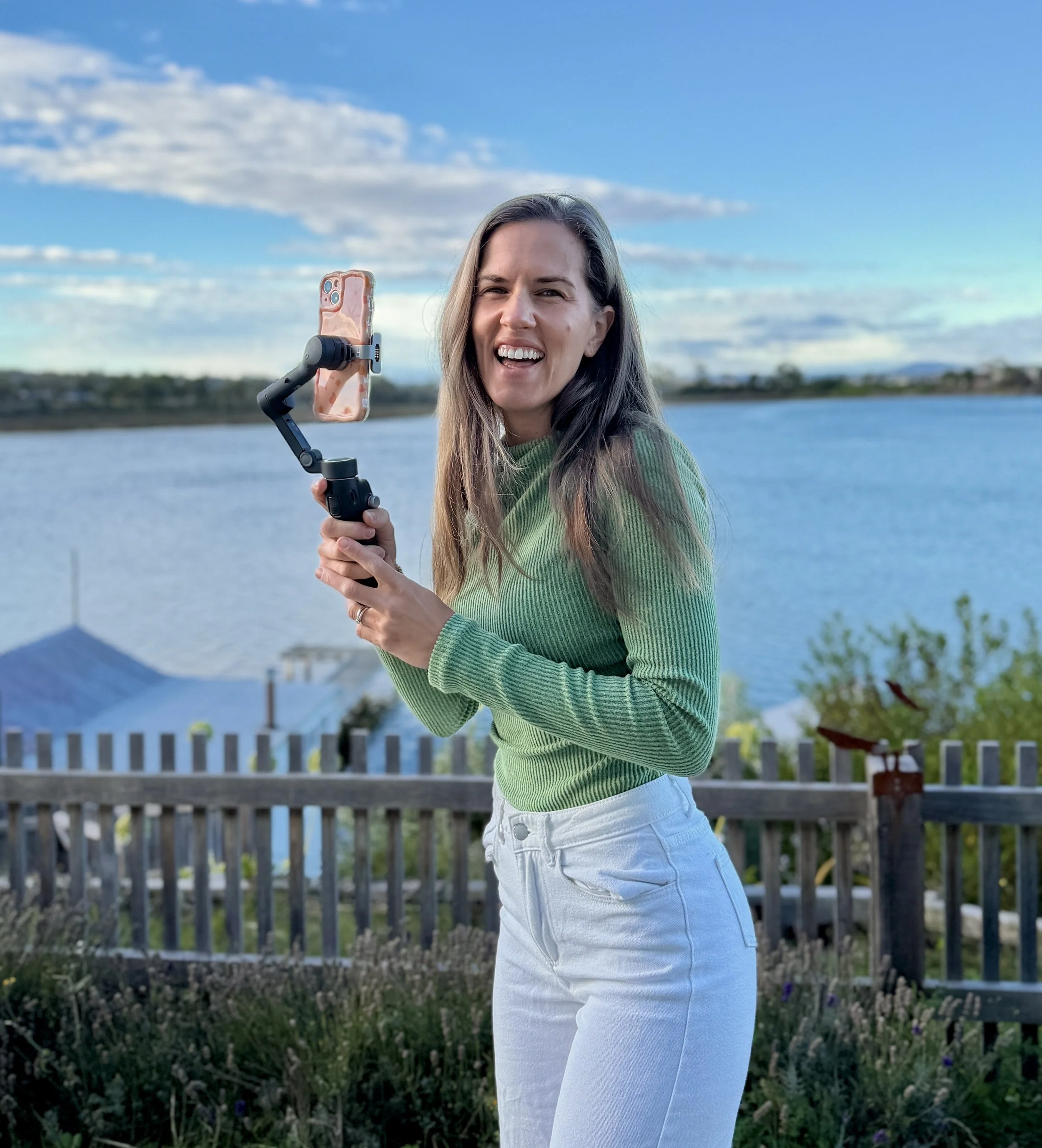 A young woman with long brown hair in a green sweater and white jeans is outdoors near a body of water, holding a selfie stick with a smartphone attached, smiling and talking.