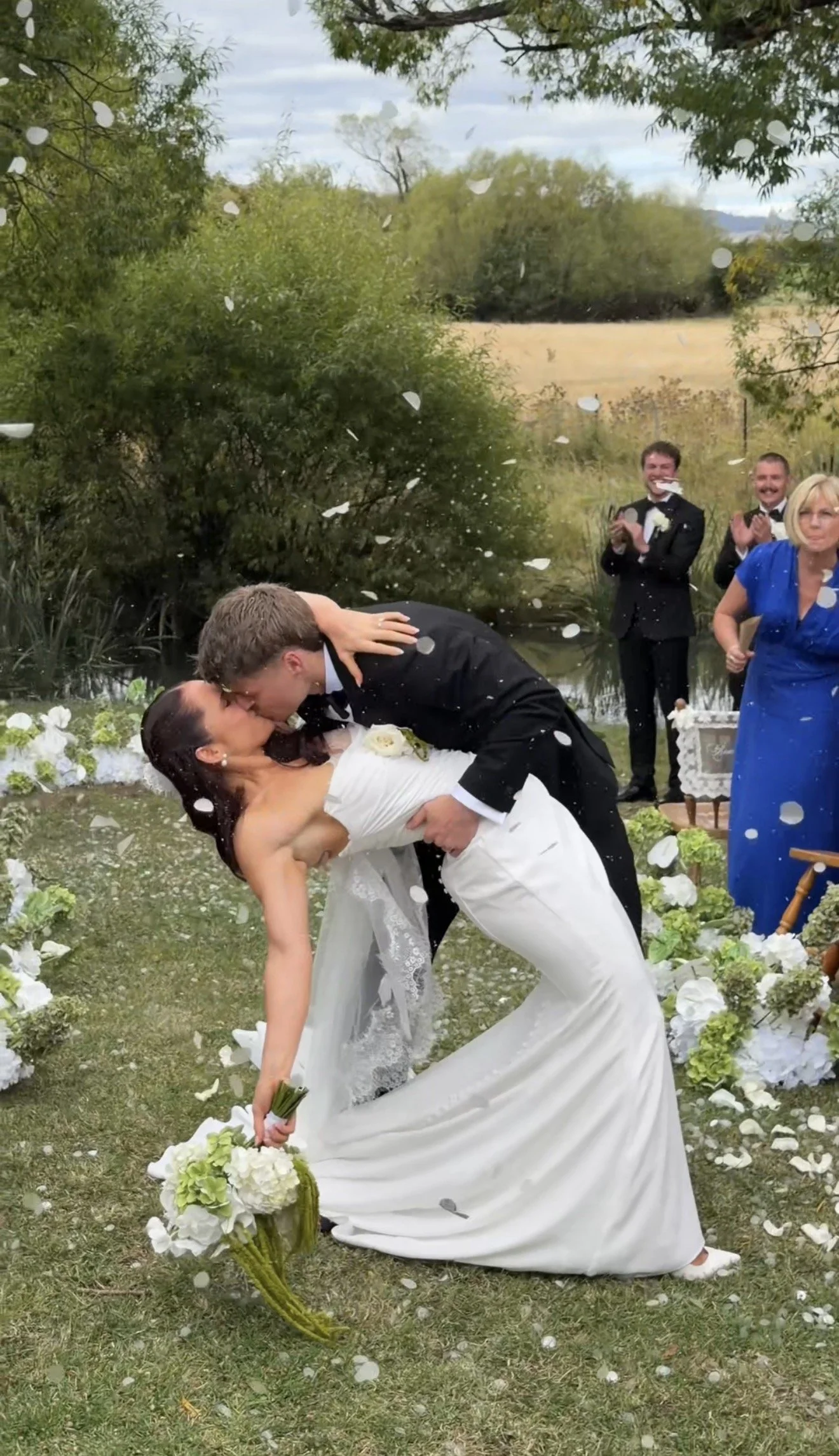 A bride and groom sharing a kiss during their outdoor wedding ceremony, with guests applauding in the background amid falling flower petals.