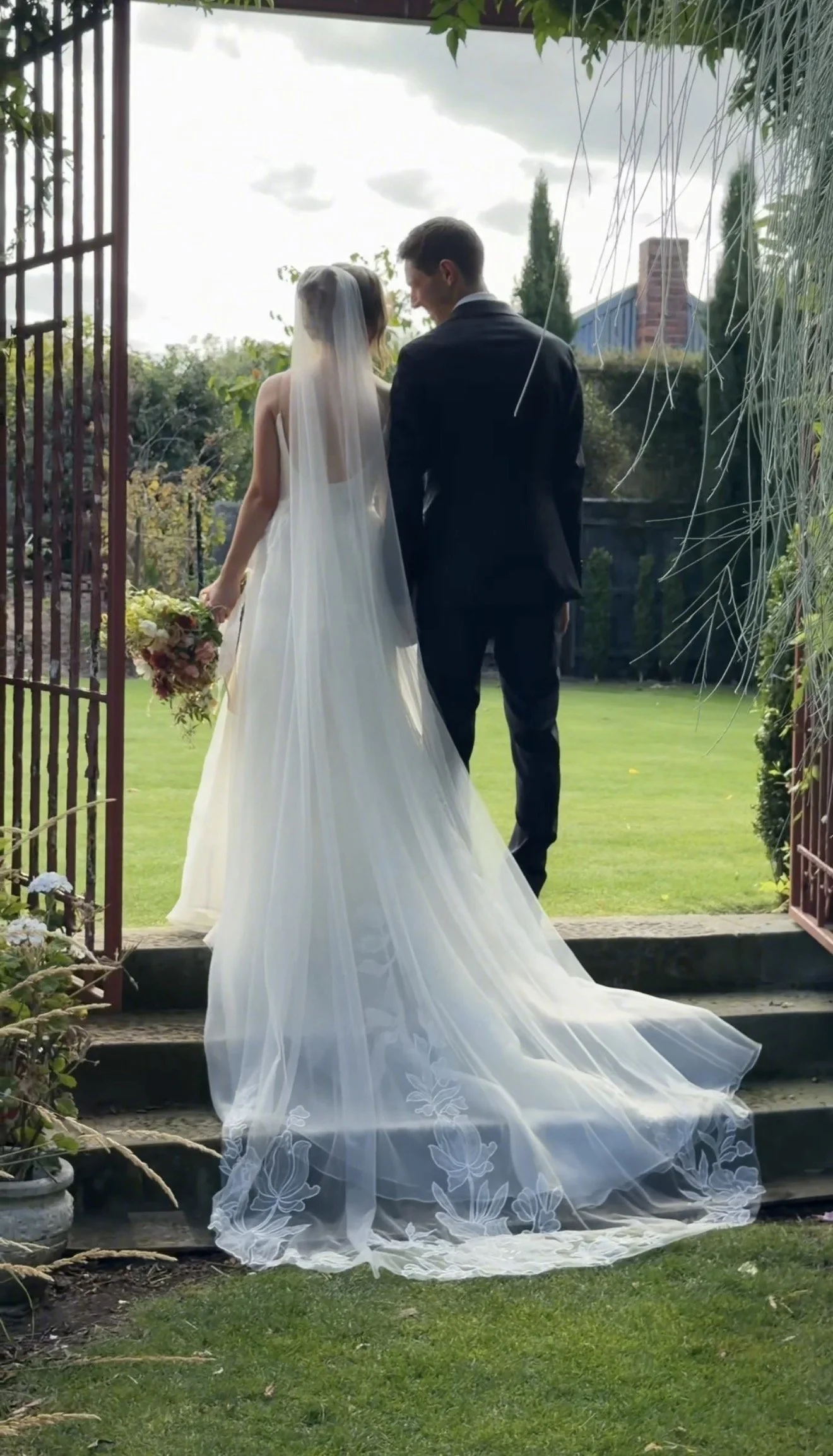 Bride and groom standing together outdoors, facing away from the camera, on a garden patio. The bride is in a white wedding gown with a dotted veil and holding a bouquet, while the groom is in a dark suit. They are surrounded by greenery, trees, and a cloudy sky.