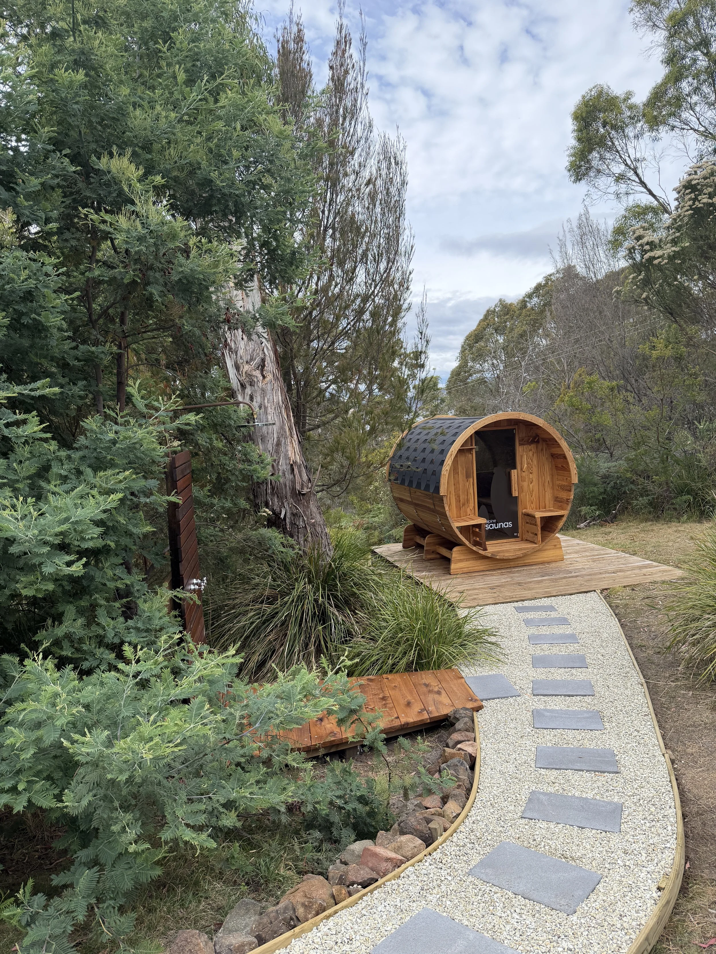 A backyard scene featuring a wooden barrel sauna, surrounding trees and plants, a stepping stone pathway, and a cloudy sky.