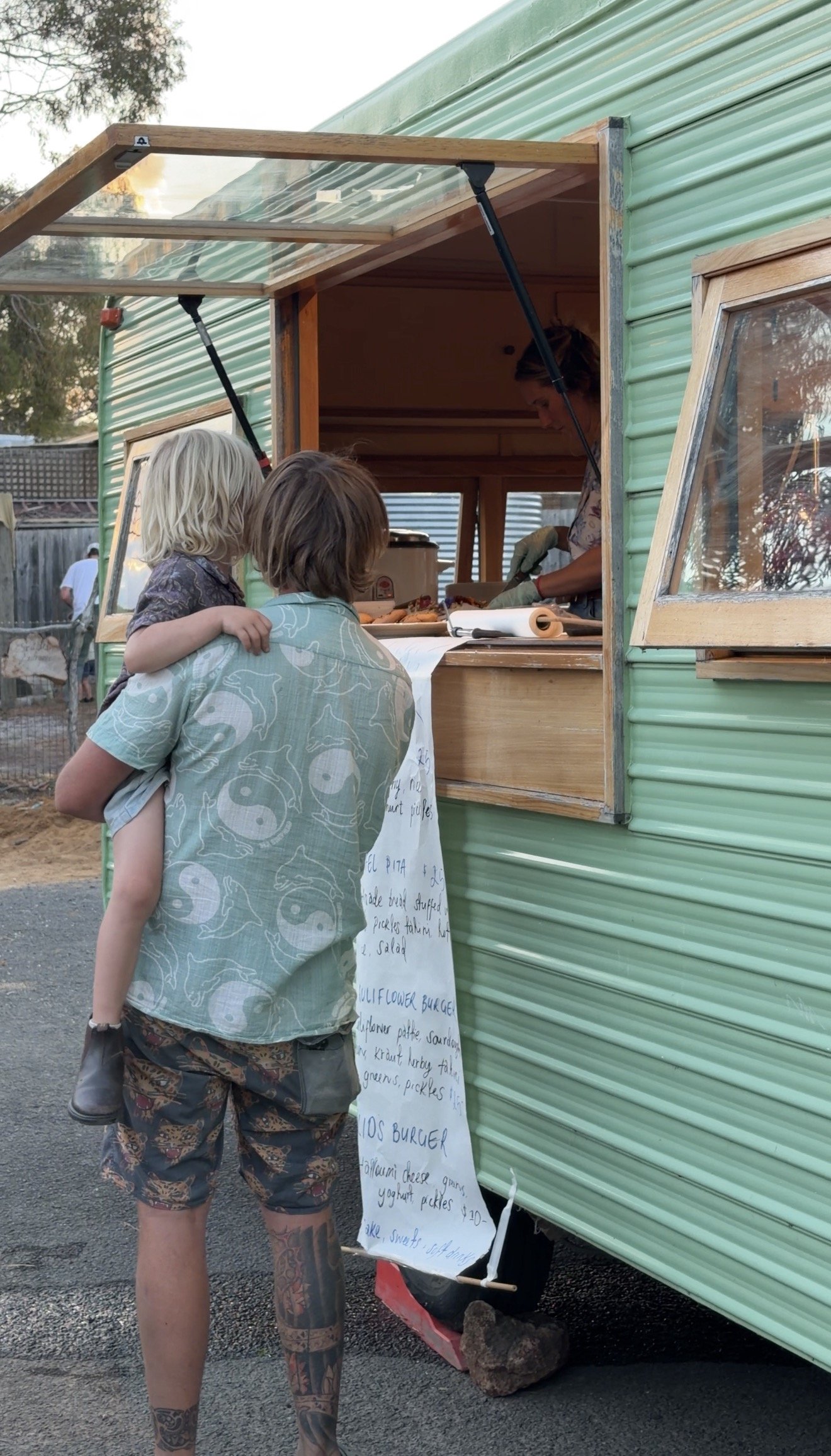 A man with tattoos on his legs and arms is standing in front of a food truck, looking at the menu written on a whiteboard. The food truck has a green exterior and a window opening for serving customers. Inside, a woman is preparing food.