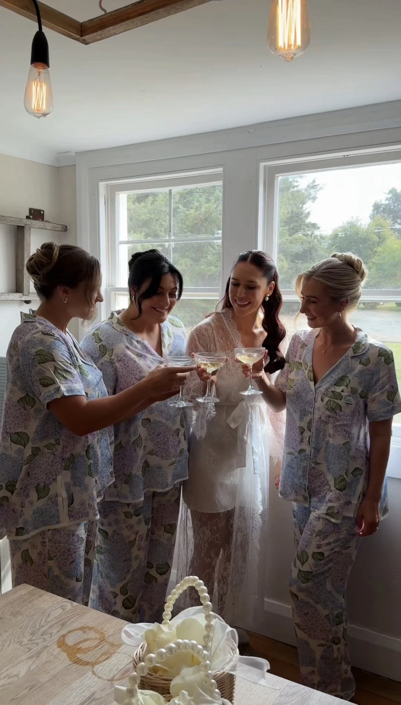 Four women celebrating, wearing matching floral pajamas, holding champagne glasses, standing near a window with greenery outside.