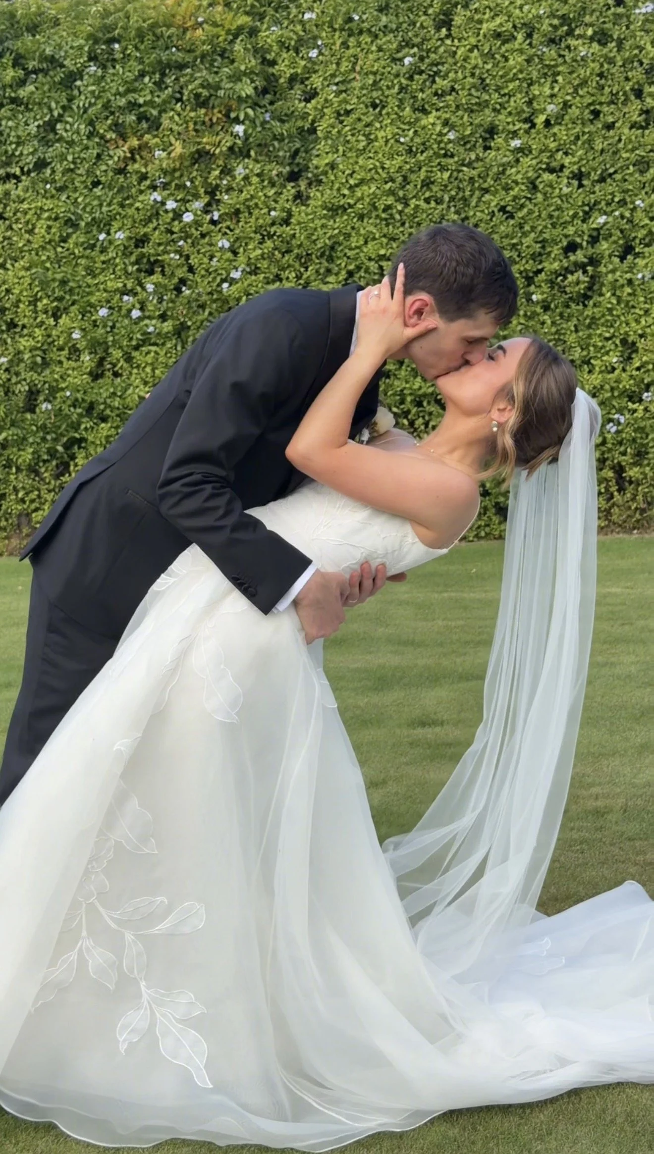 A bride and groom sharing a kiss outdoors on a grassy lawn with a green hedge in the background. The groom is in a black tuxedo, and the bride is in a white wedding gown with a long veil.