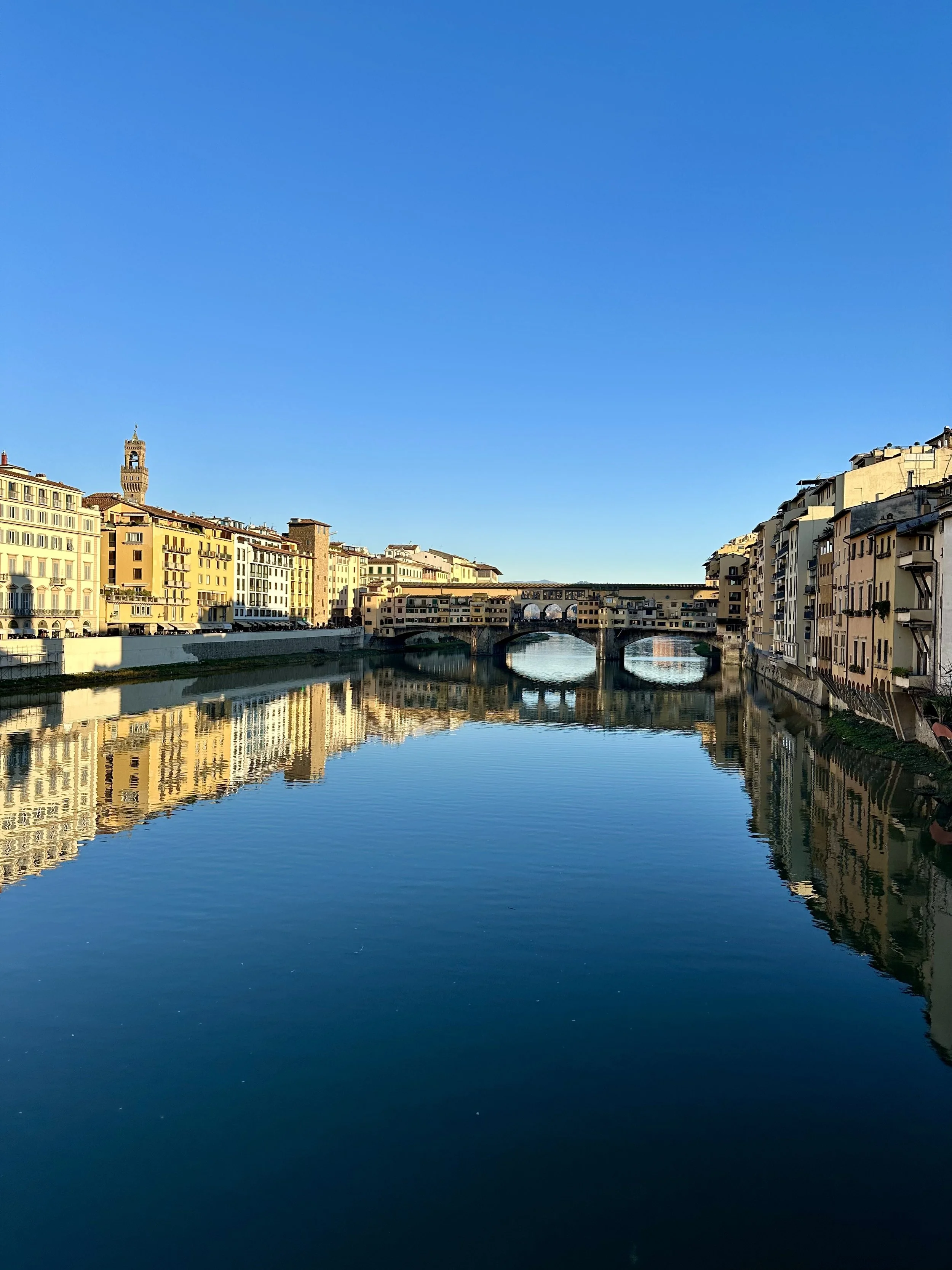 Sky above a city with buildings along a river, reflecting in the water, with a bridge and a tower visible in the background.