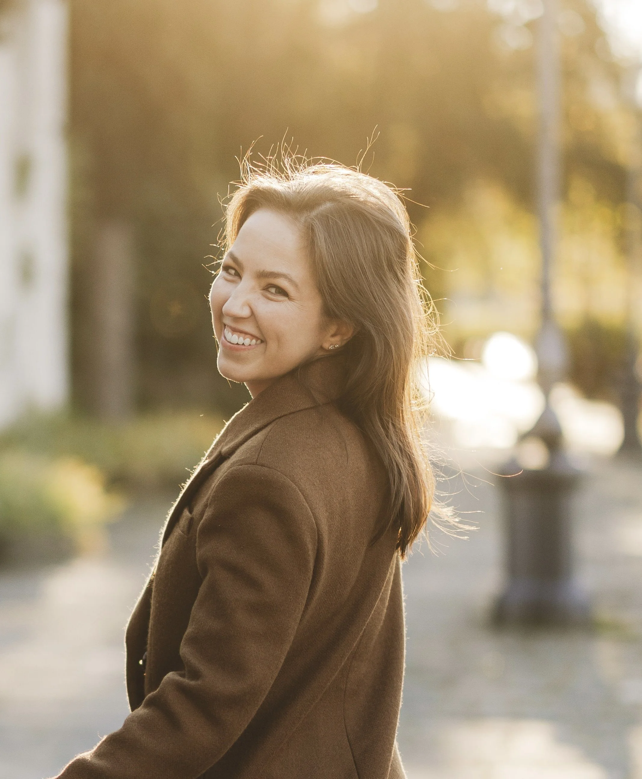 Portrait of a smiling woman with long brown hair, wearing a brown coat, outdoors in warm sunlight with blurred background of trees and street.