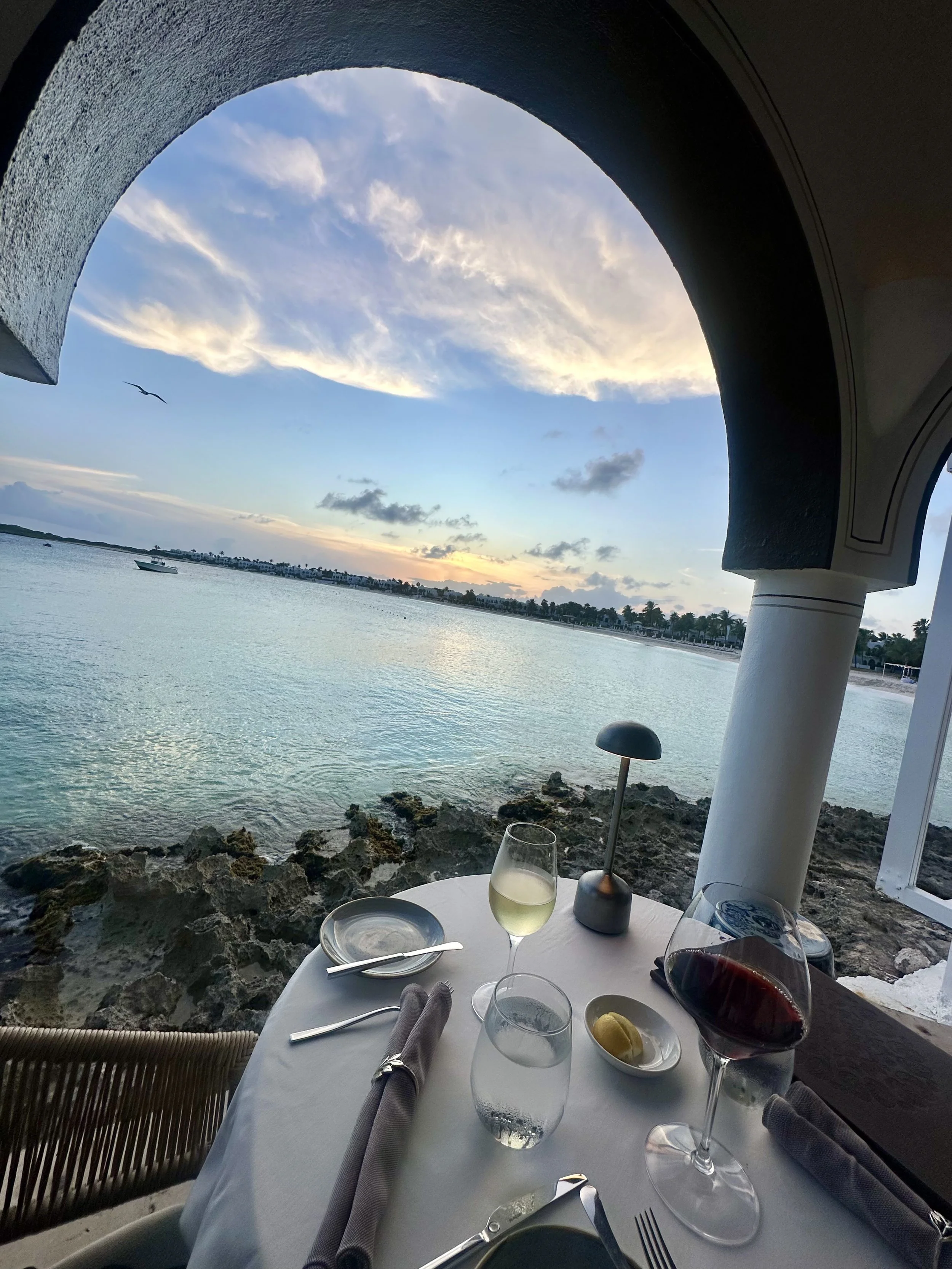 A table set with wine glasses, a plate of food, and utensils overlooking a body of water and a cloudy sky at sunset, viewed from a balcony or patio.