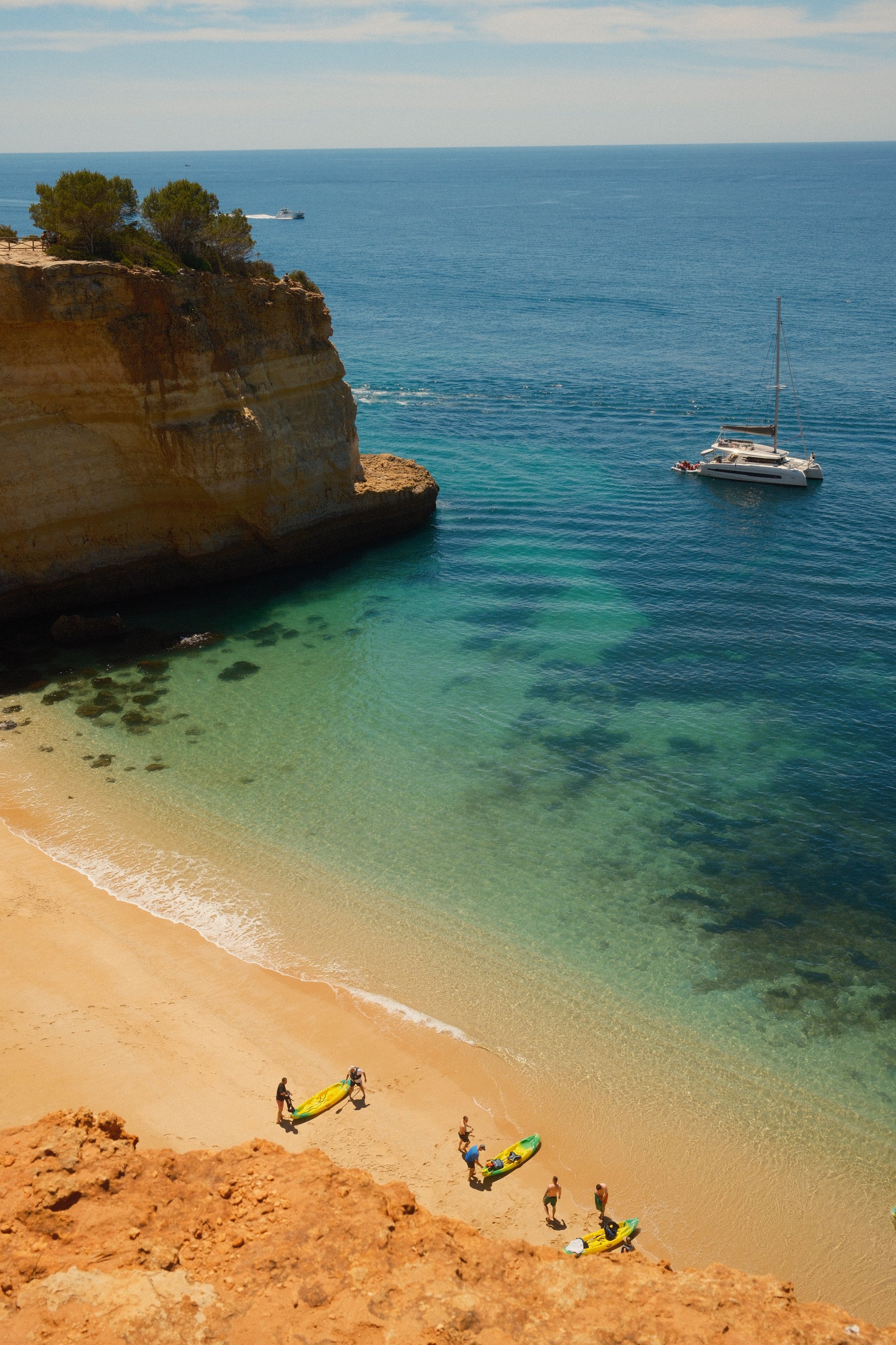 A coastal scene with a sandy beach, clear turquoise water, and a rocky cliff with vegetation. People are preparing kayaks on the beach. A sailboat floats on the water, and a boat is seen in the distance.