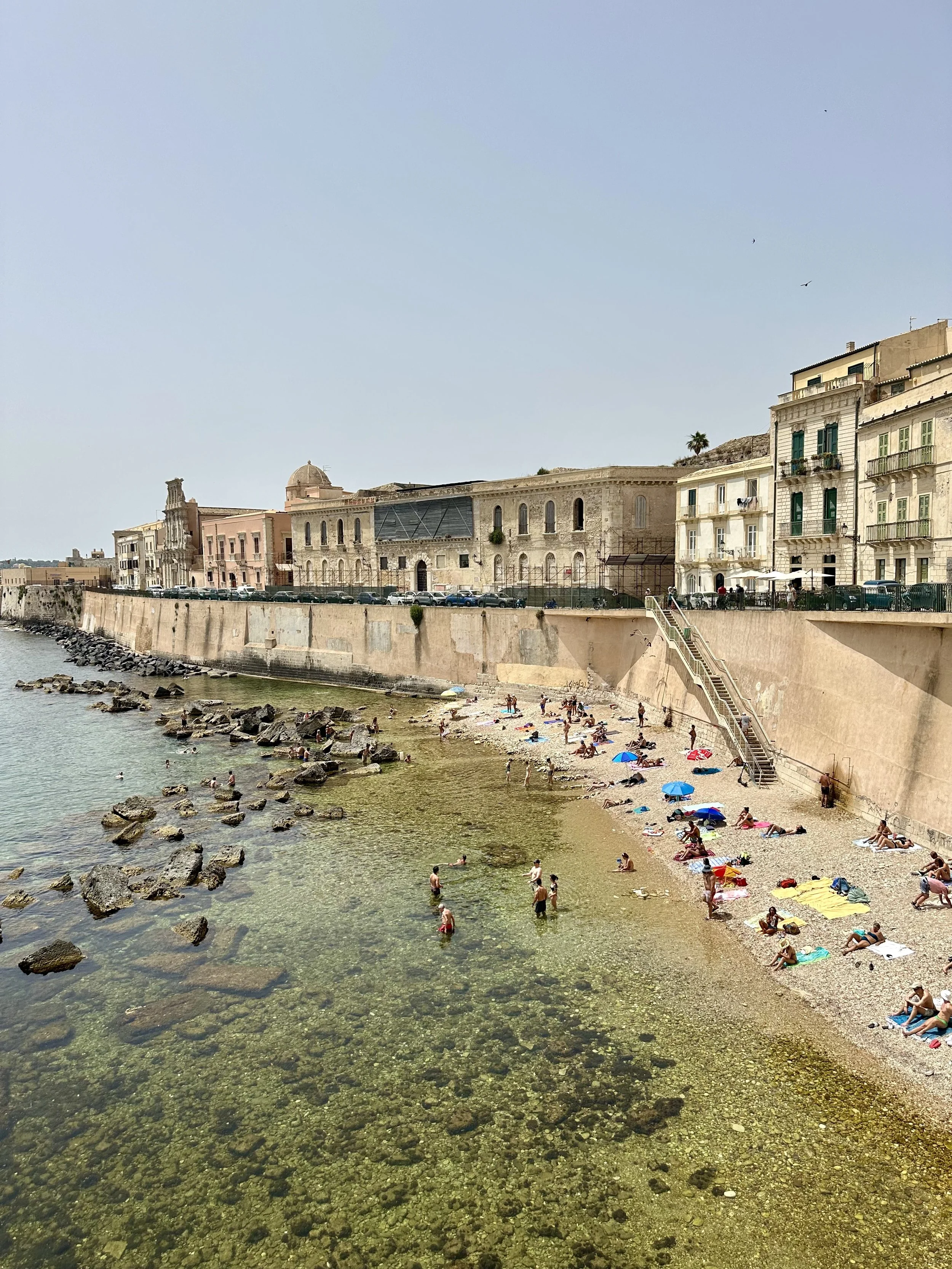 View of a small beach with many people relaxing on towels and umbrellas, along a rocky shoreline with clear water, and historic buildings in the background.
