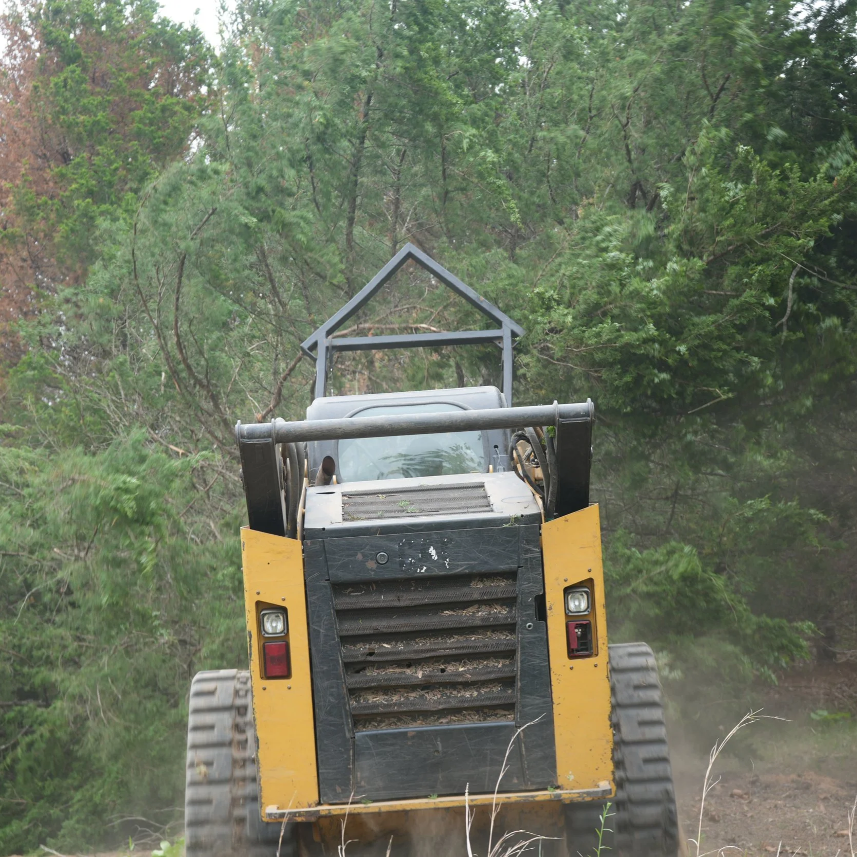 A small yellow bulldozer moving dirt or debris among green trees in a wooded area.