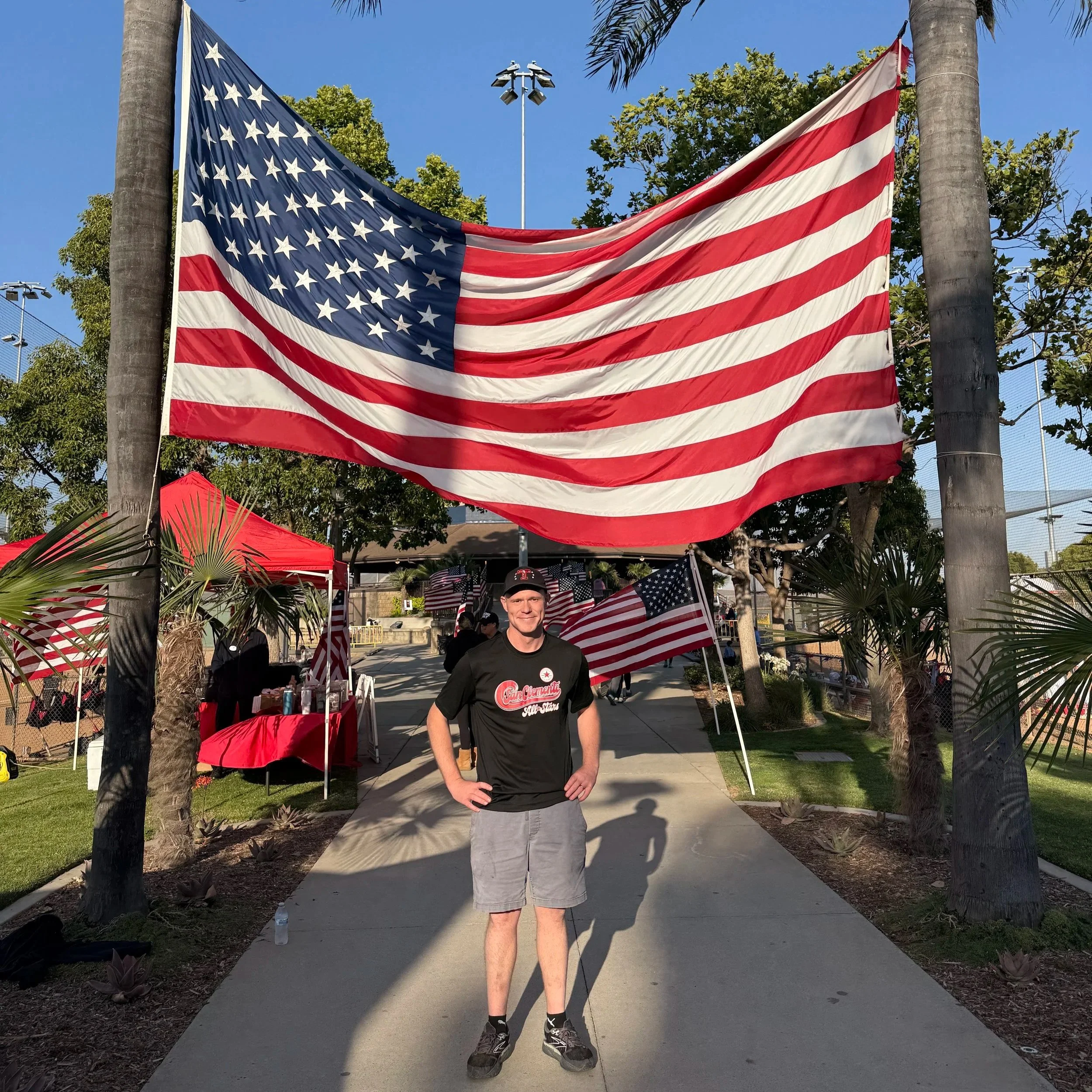 A young man wearing a black shirt with a sports logo and khaki shorts standing on a sidewalk in front of an American flag hanging between two trees on a sunny day.