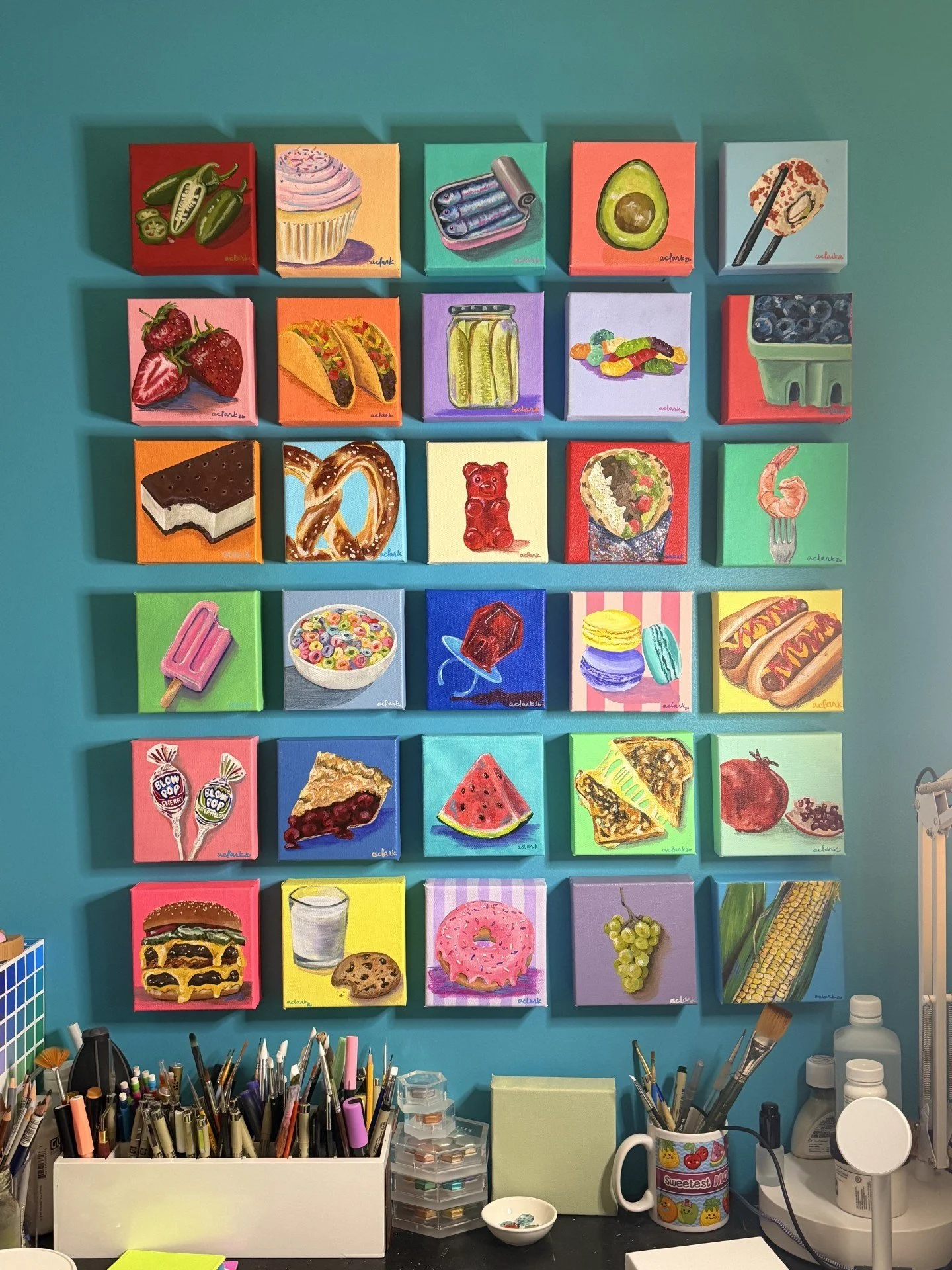 Tastes of Joy mini food paintings arranged in a studio grid above brushes and art supplies
