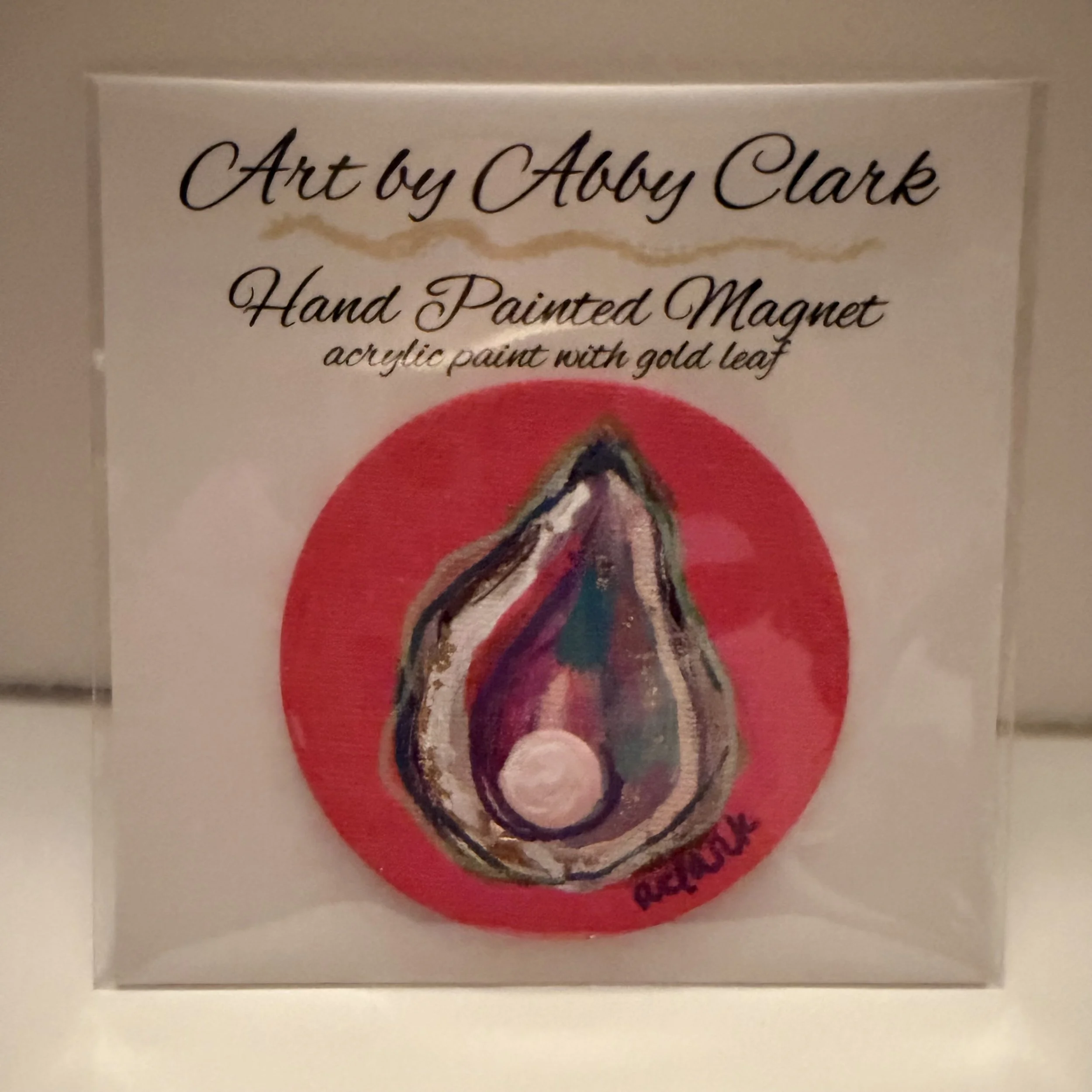 Round oyster magnet with a raised pearl embellishment against a coral-pink background, in clear packaging