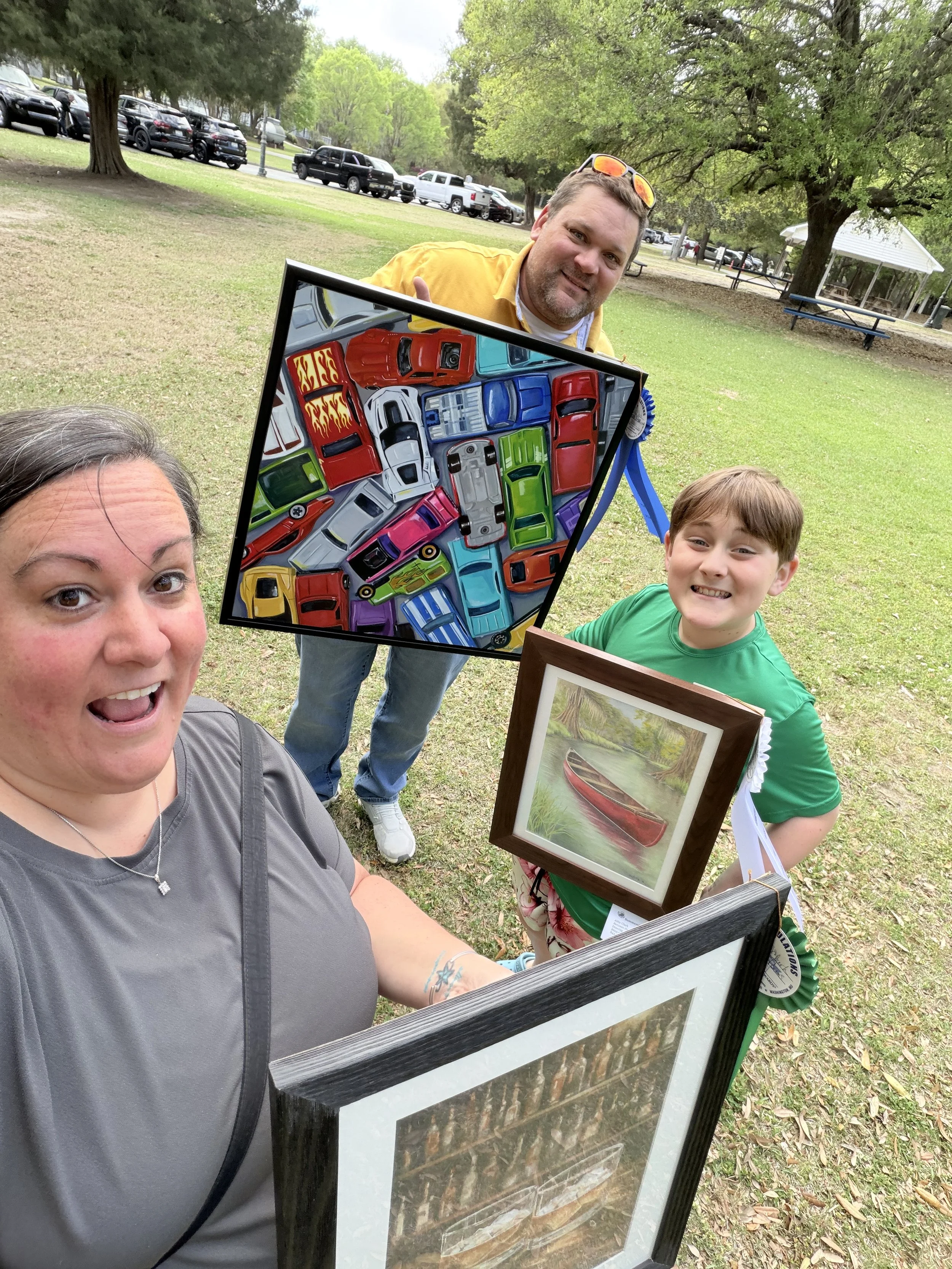 Abby Clark outdoors with framed paintings, including colorful cars and a canoe on quiet water