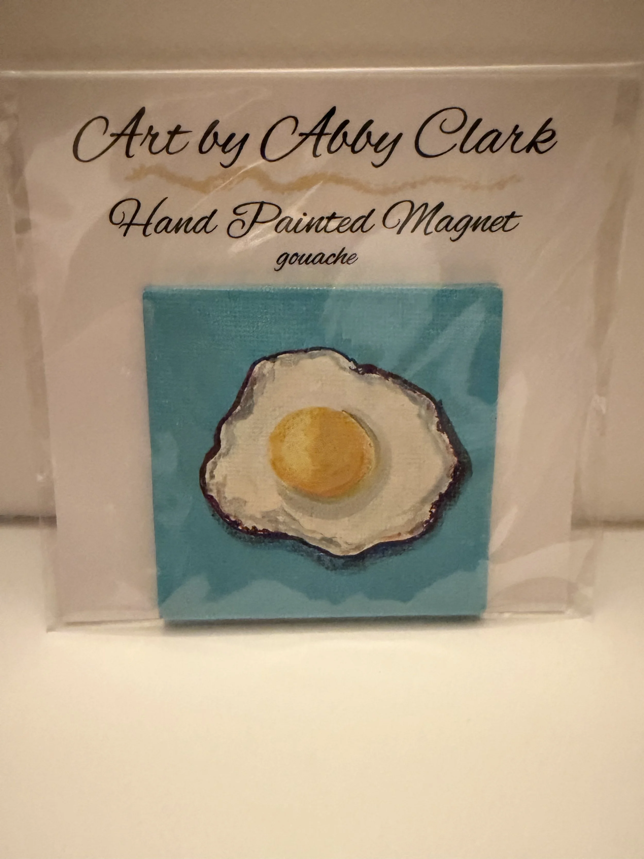 Fried egg magnet with a yellow yolk and white egg against turquoise, shown in its packaging