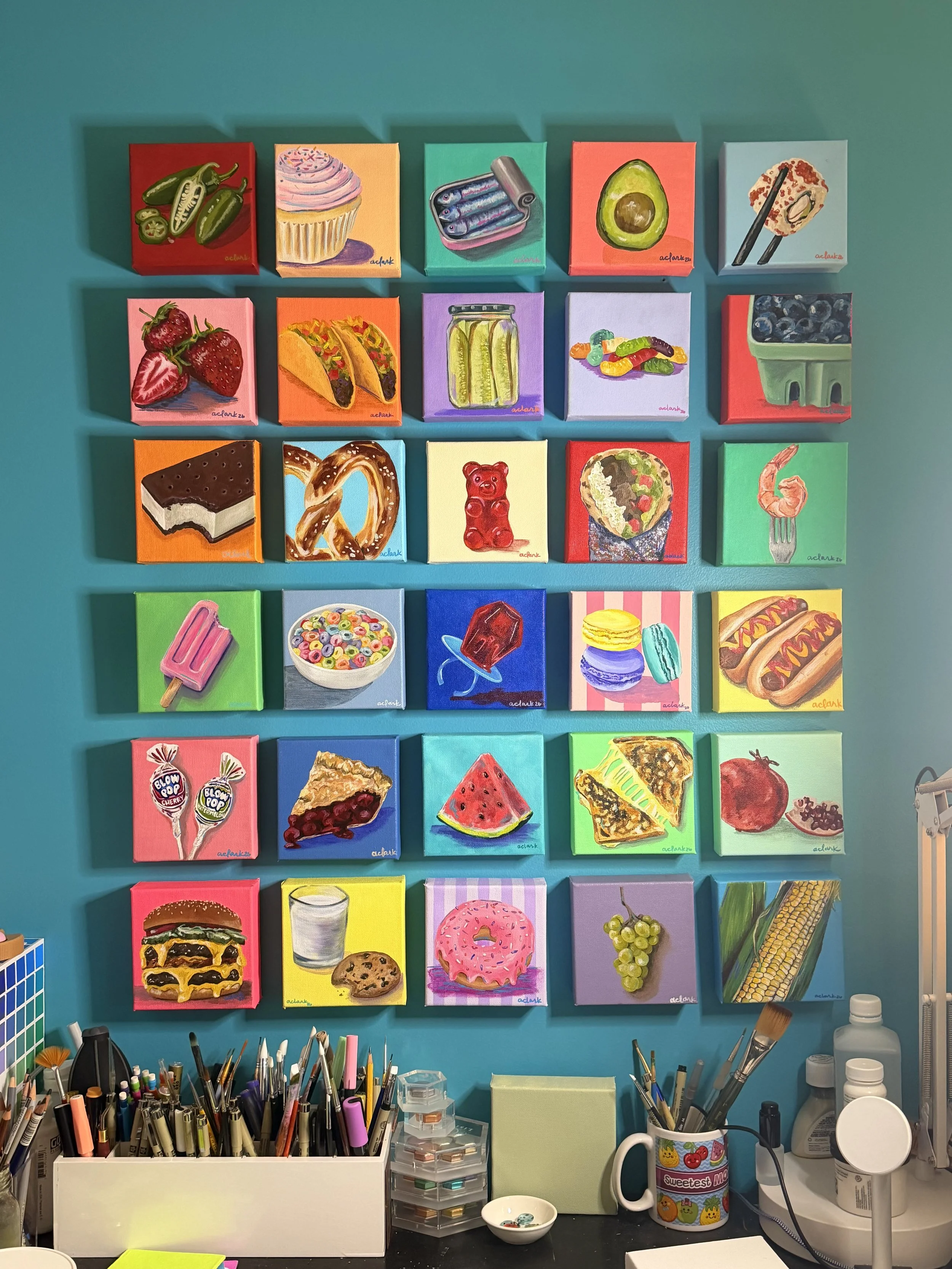 Tastes of Joy mini food paintings arranged in a studio grid above brushes and art supplies