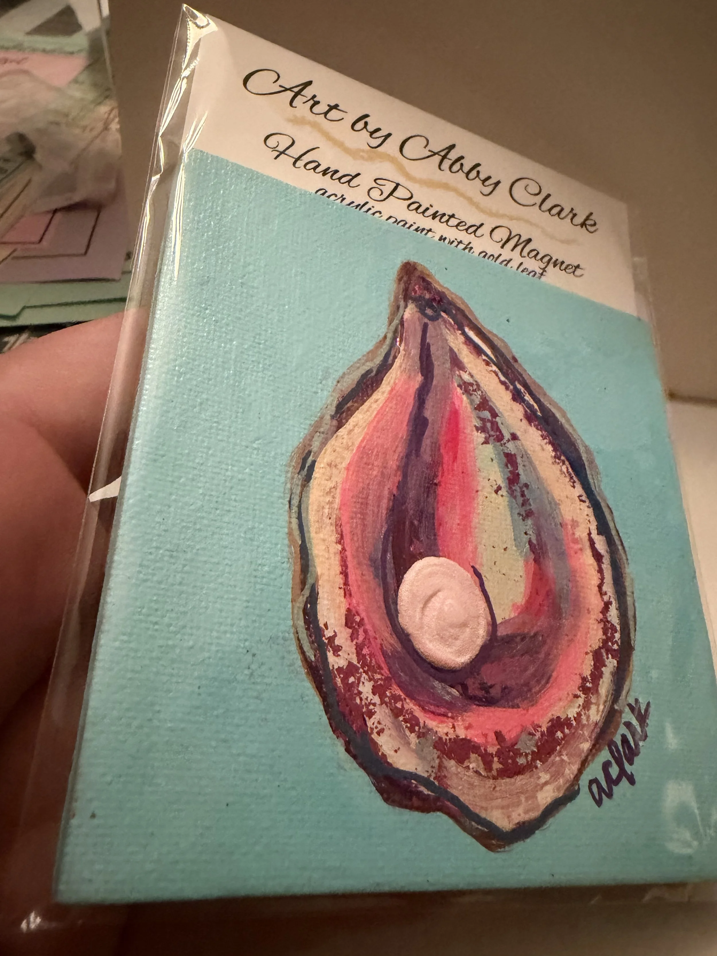 Angled view of Abby Clark’s hand-painted oyster canvas magnet, showing metallic accents, a raised pearl and turquoise background