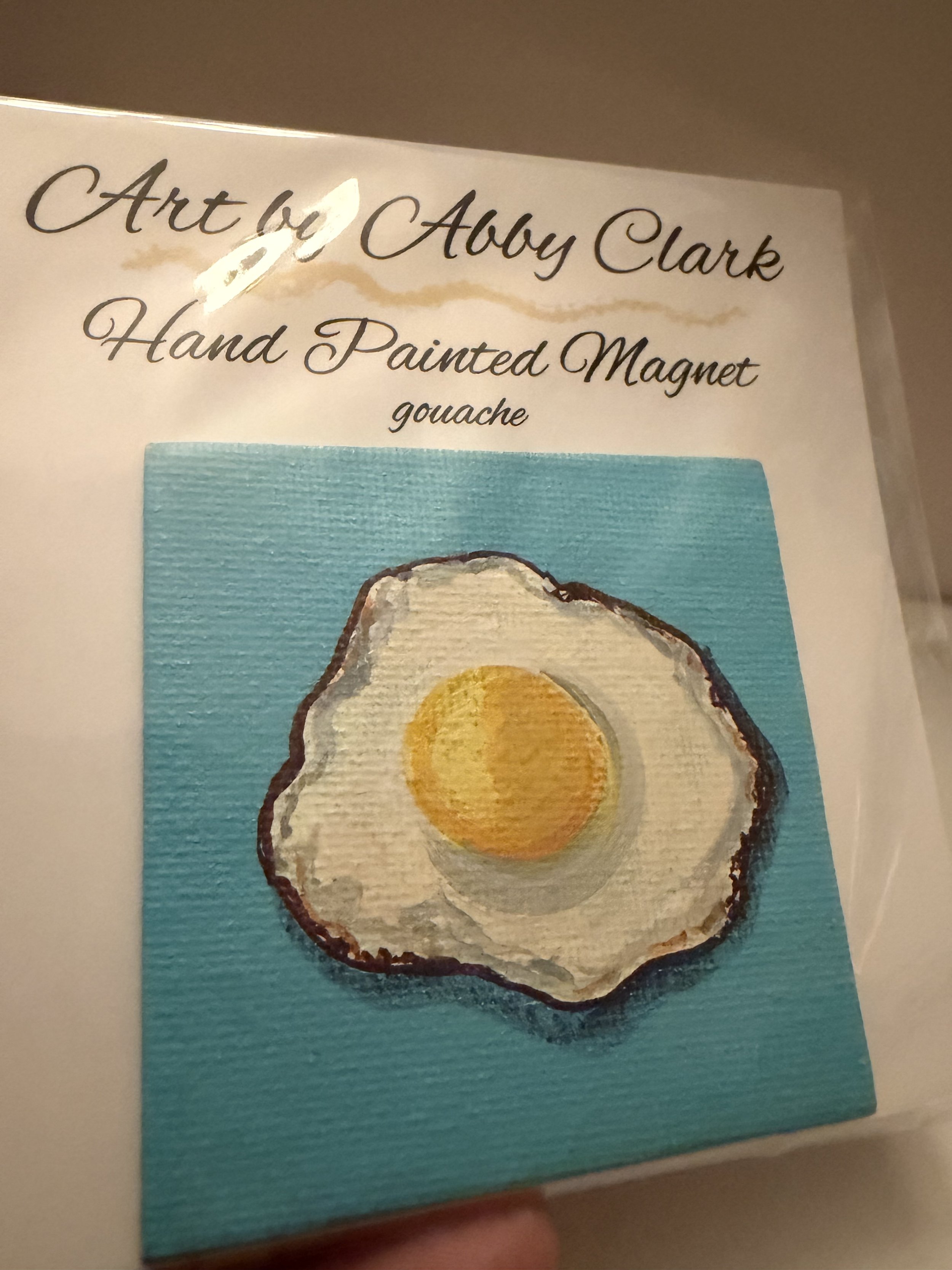 Angled view of Abby Clark’s fried egg magnet painted on a turquoise mini canvas, in clear packaging