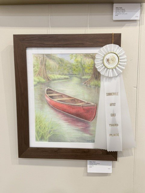 Between the Shores displayed in a brown frame with a white mat and an award ribbon beside the artwork