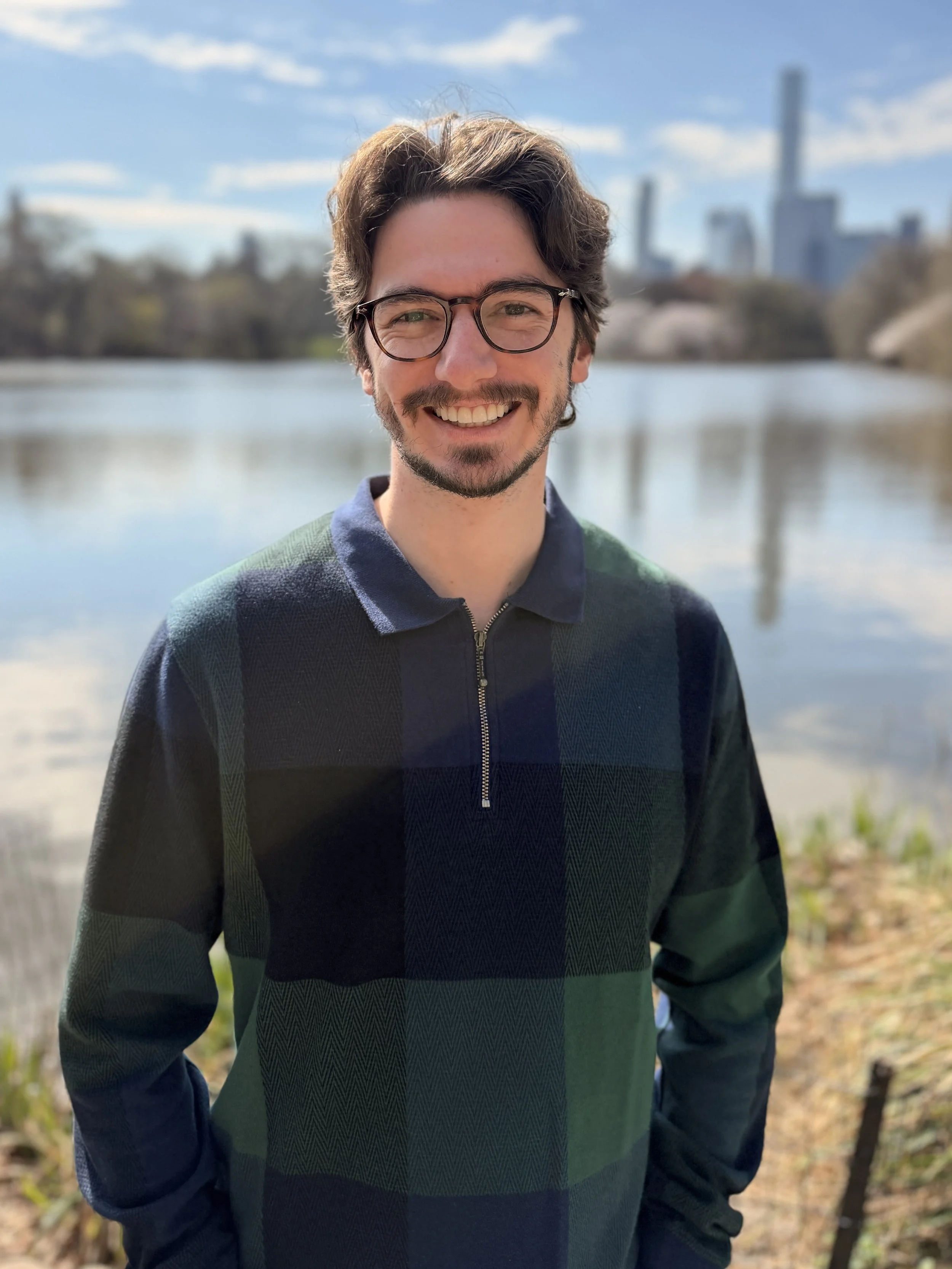 A smiling man with glasses and a beard standing outdoors near a body of water, with city skyscrapers in the background on a sunny day.