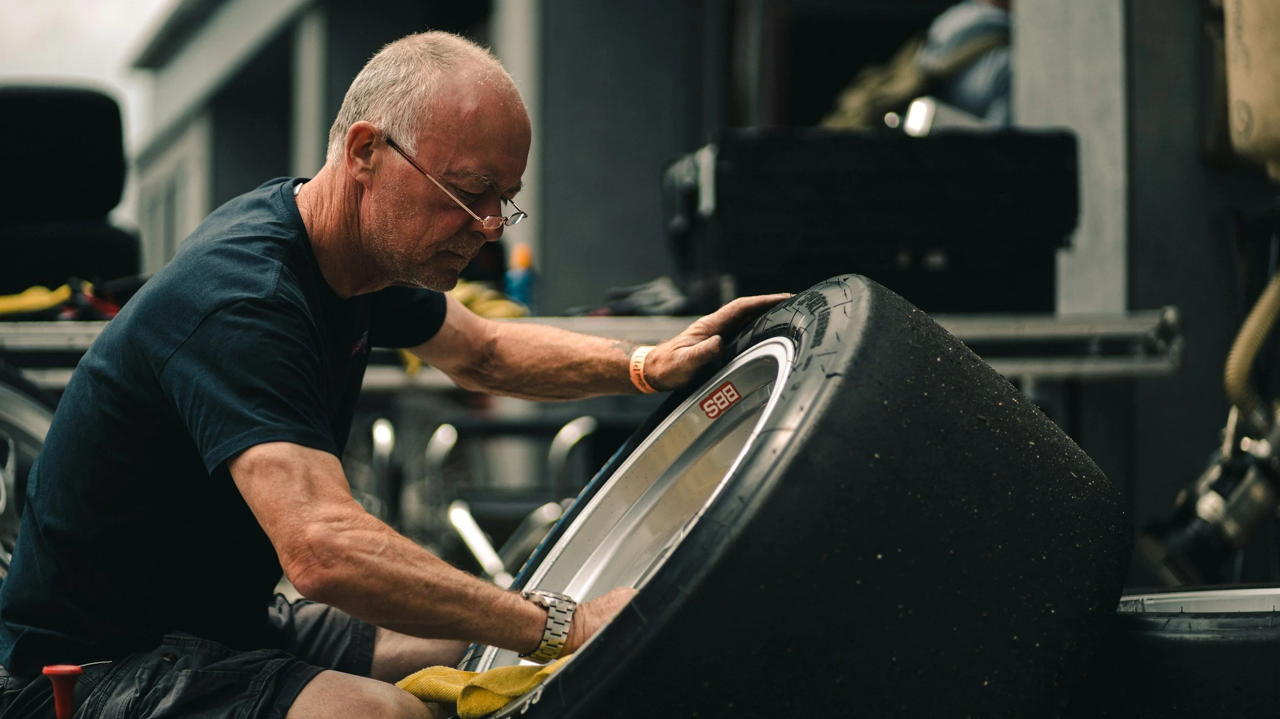 An older man with glasses working on a large car tire in a workshop.