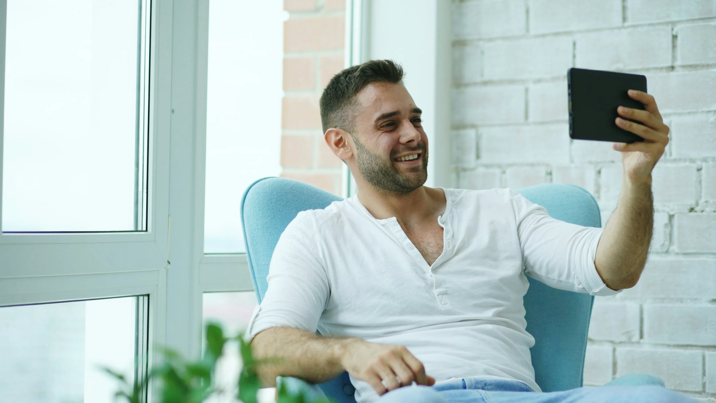 A man sitting on a light blue chair, smiling and performing a video call, near a window with a white brick wall background.