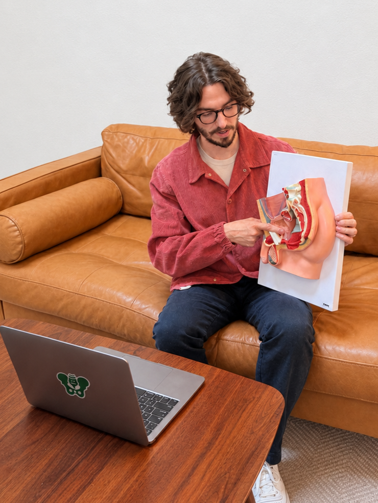 A man with curly hair and glasses sitting on a tan leather couch, pointing at a model of human anatomy held in his other hand, with a laptop on the table in front of him.