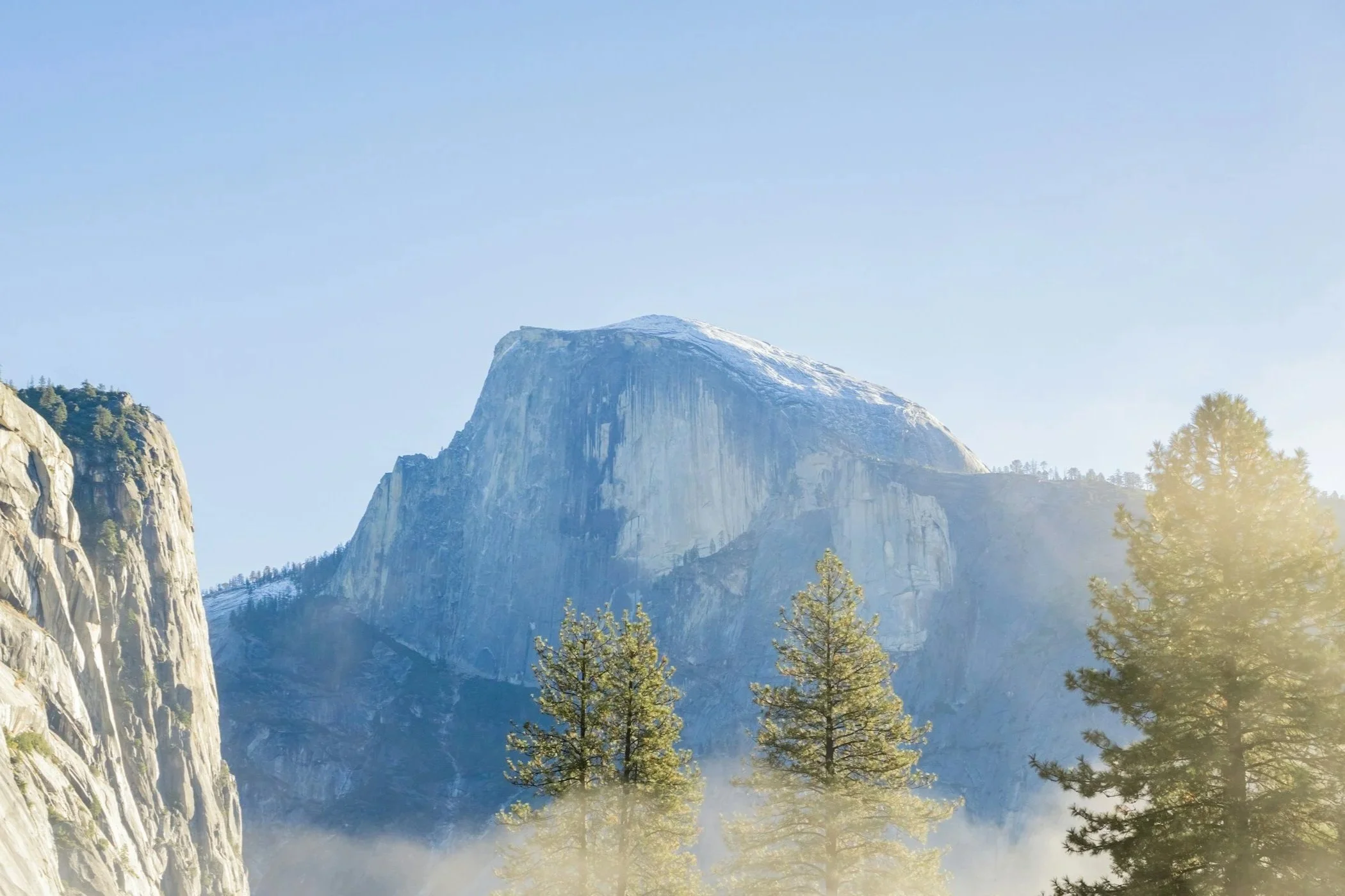 A mountain landscape with a prominent granite peak, surrounded by forested areas and a clear blue sky.