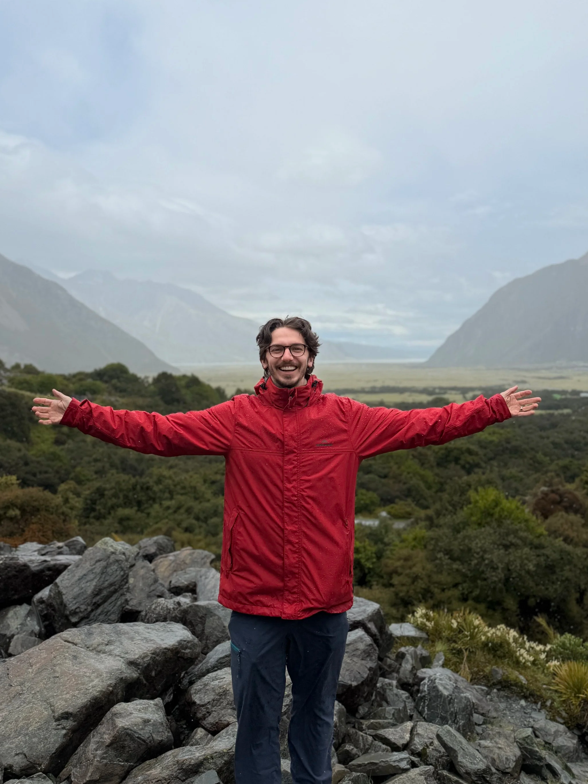 A man in a red jacket standing on rocks in a mountainous landscape with greenery and clouds in the sky, arms outstretched and smiling.