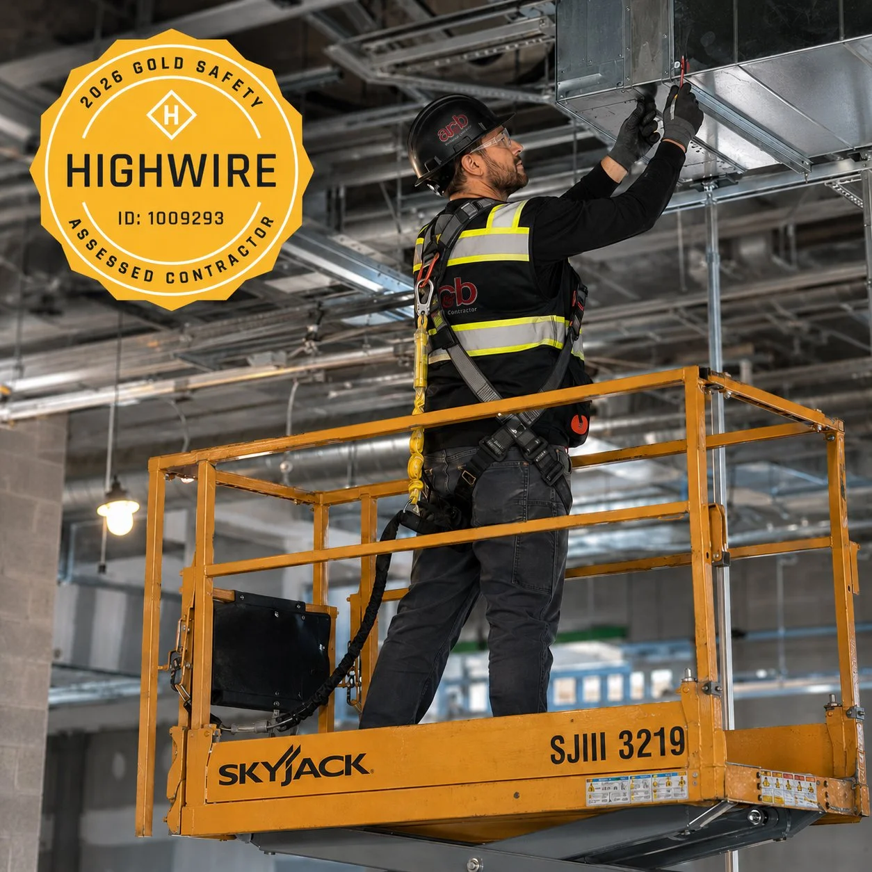 A construction worker wearing a black helmet, safety glasses, and a reflective vest stands on an orange lift platform working on an electrical or HVAC installation on the ceiling of a building under construction.